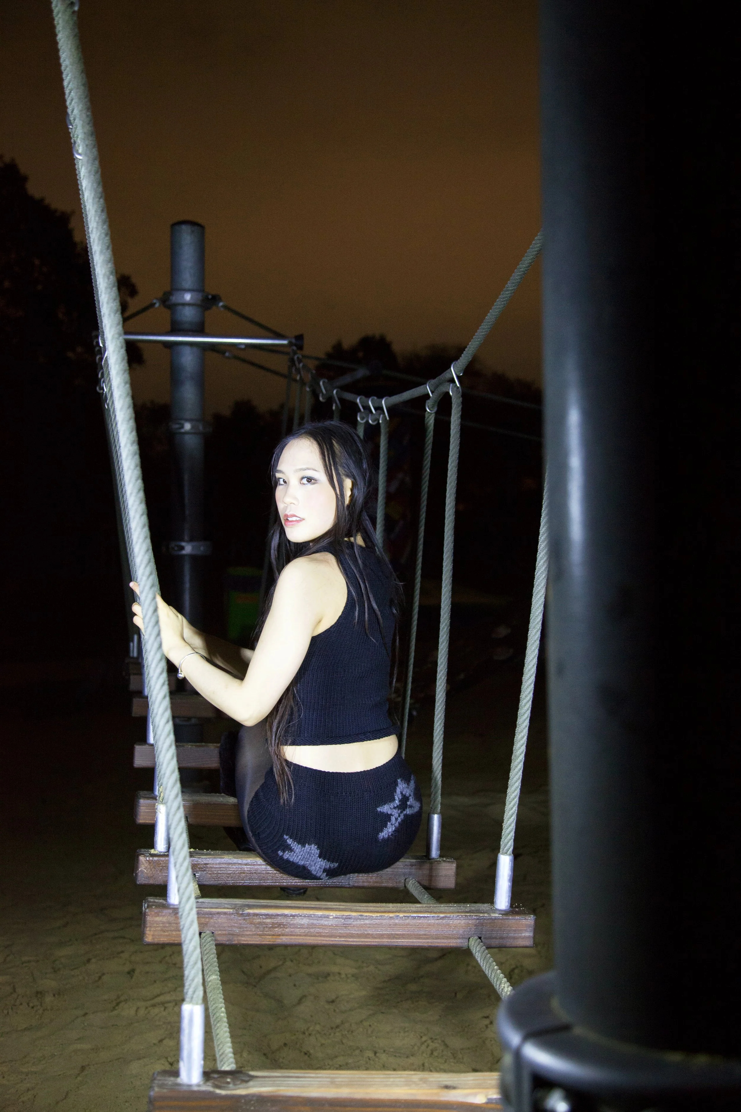 A young woman with long dark hair sitting on a swing at night, illuminated by a strong light source, with a dark sky and trees in the background.