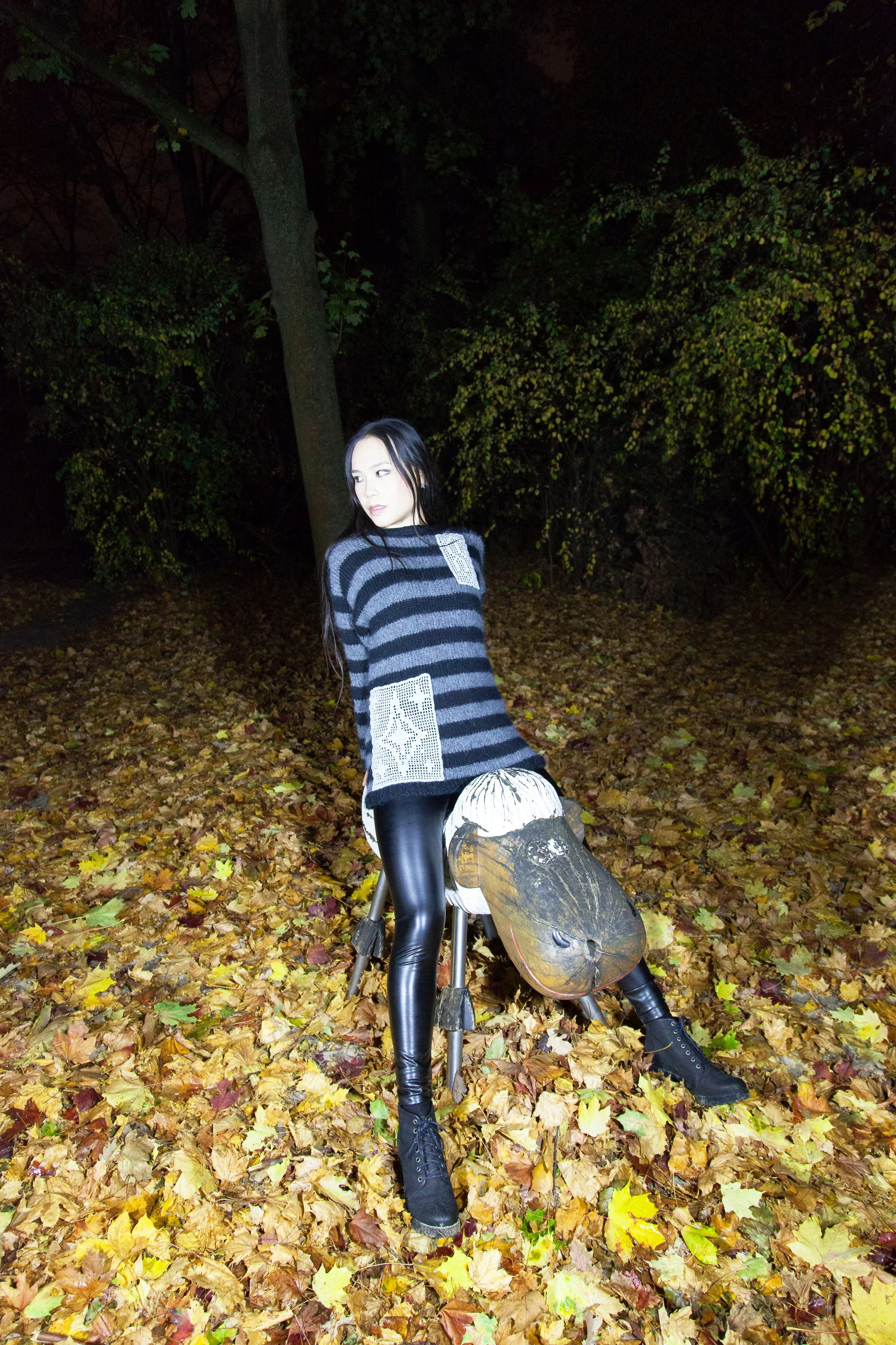 A woman with long dark hair in a striped sweater and shiny black pants poses outdoors at night, sitting on a small sheep sculpture with fallen autumn leaves and trees in the background.