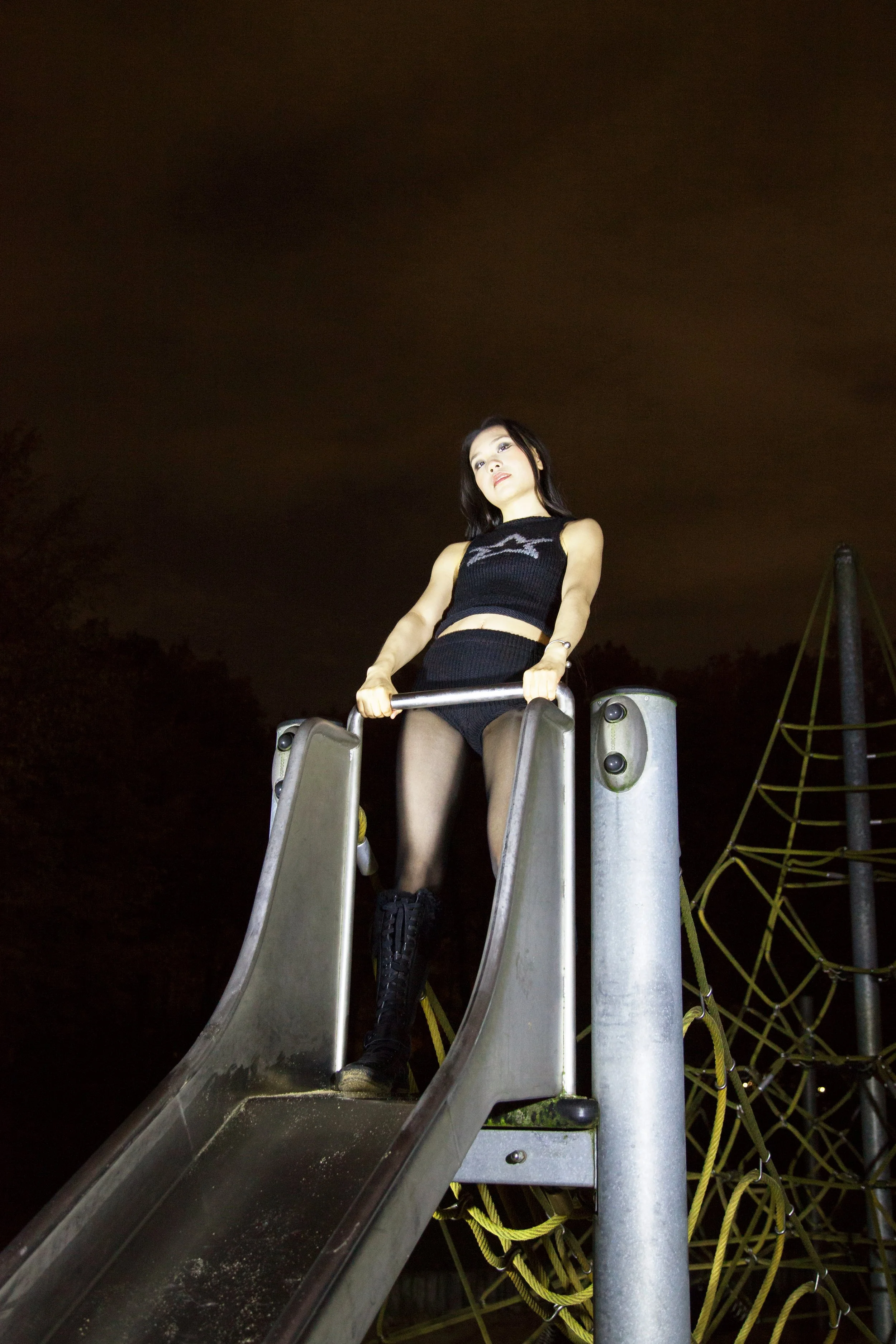 A young woman with black hair in a black crop top and shorts standing at the top of a slide in a playground at night, illuminated by a bright light.