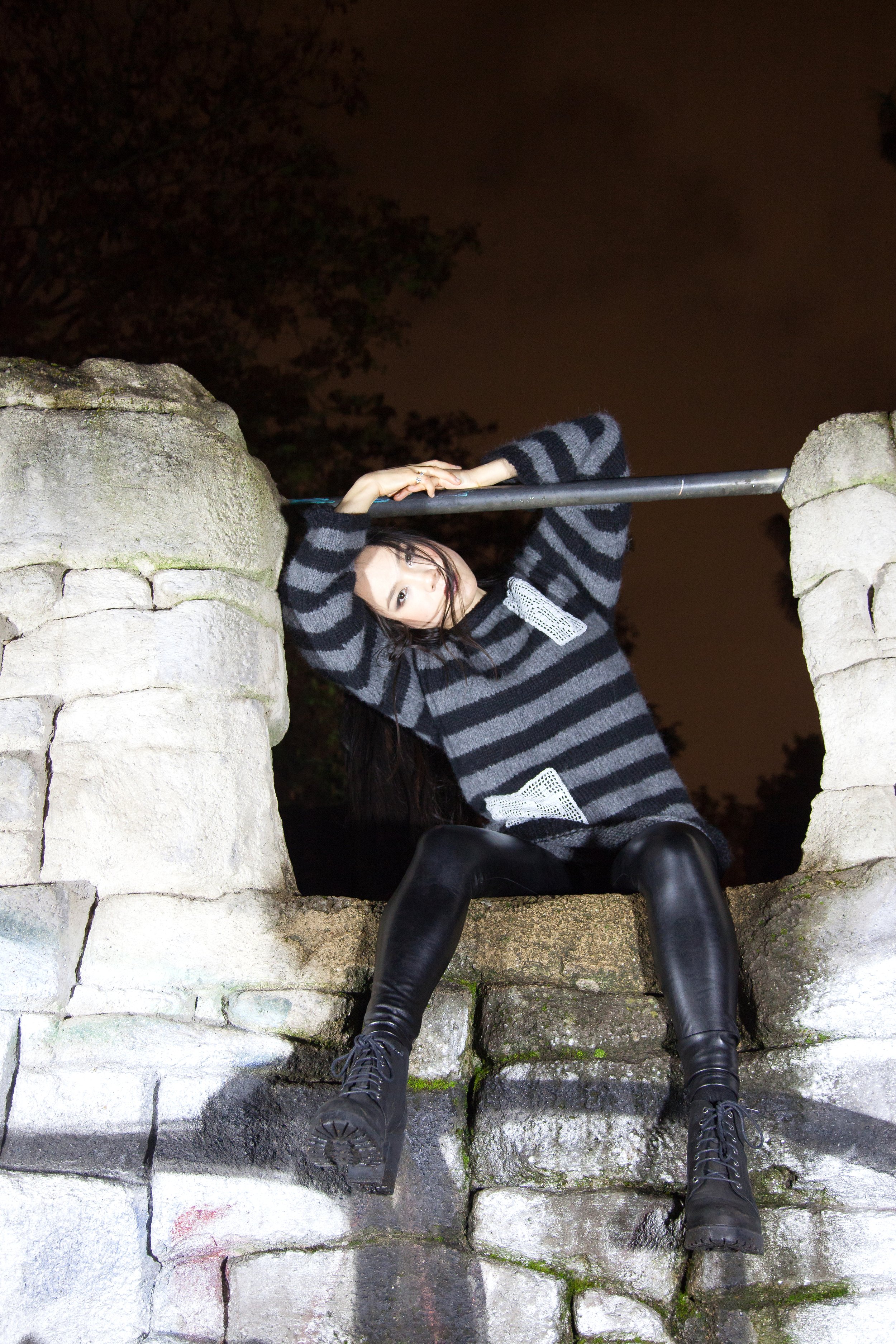 A woman with long dark hair sitting on a stone ledge at night, holding onto a metal pole with one hand, wearing a black and gray striped sweater, black leather pants, and black platform boots.