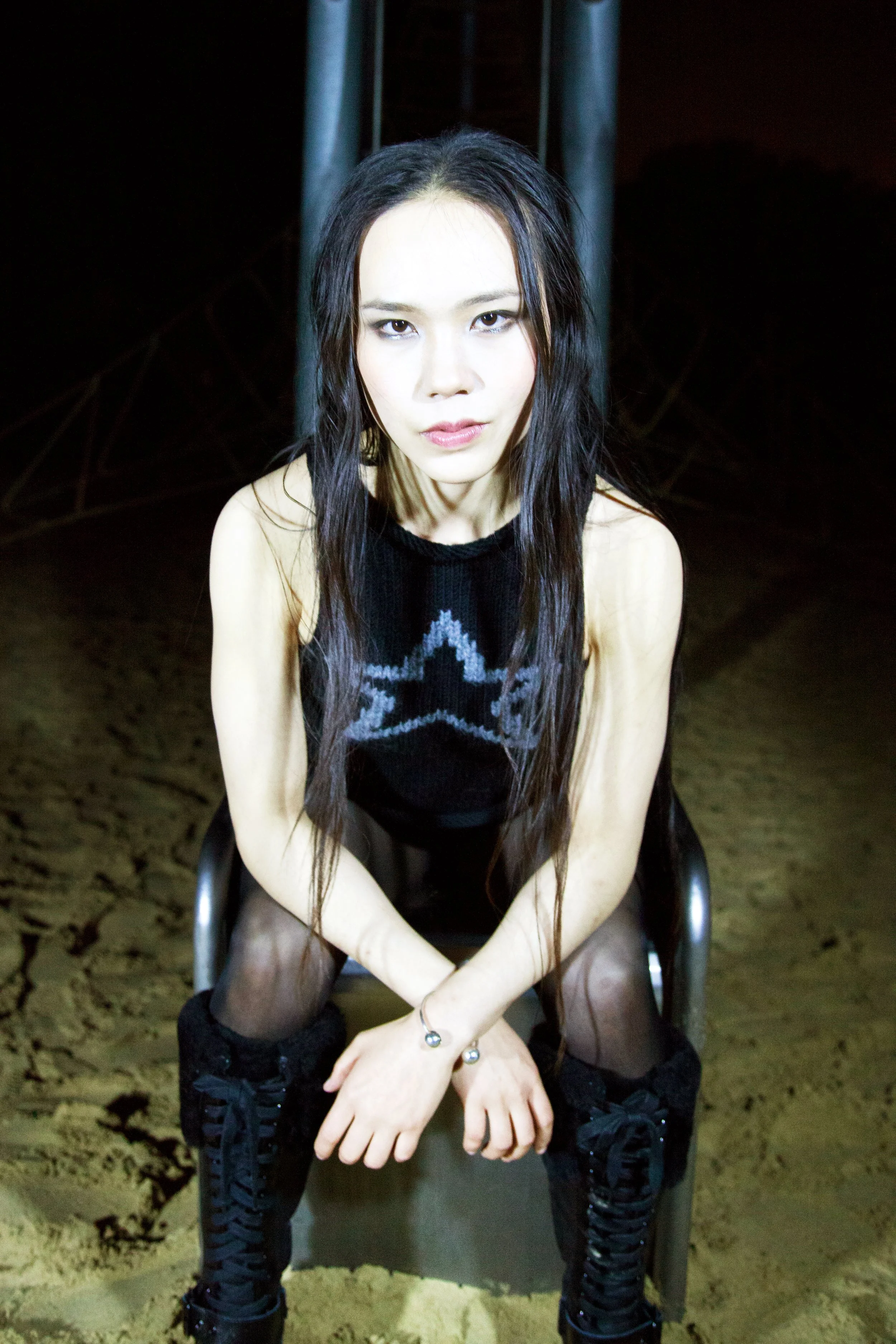 A young woman with long black hair sitting on a metal slide in a dark, sandy area, wearing a sleeveless black sweater with a grey star pattern, black tights, and black lace-up boots.