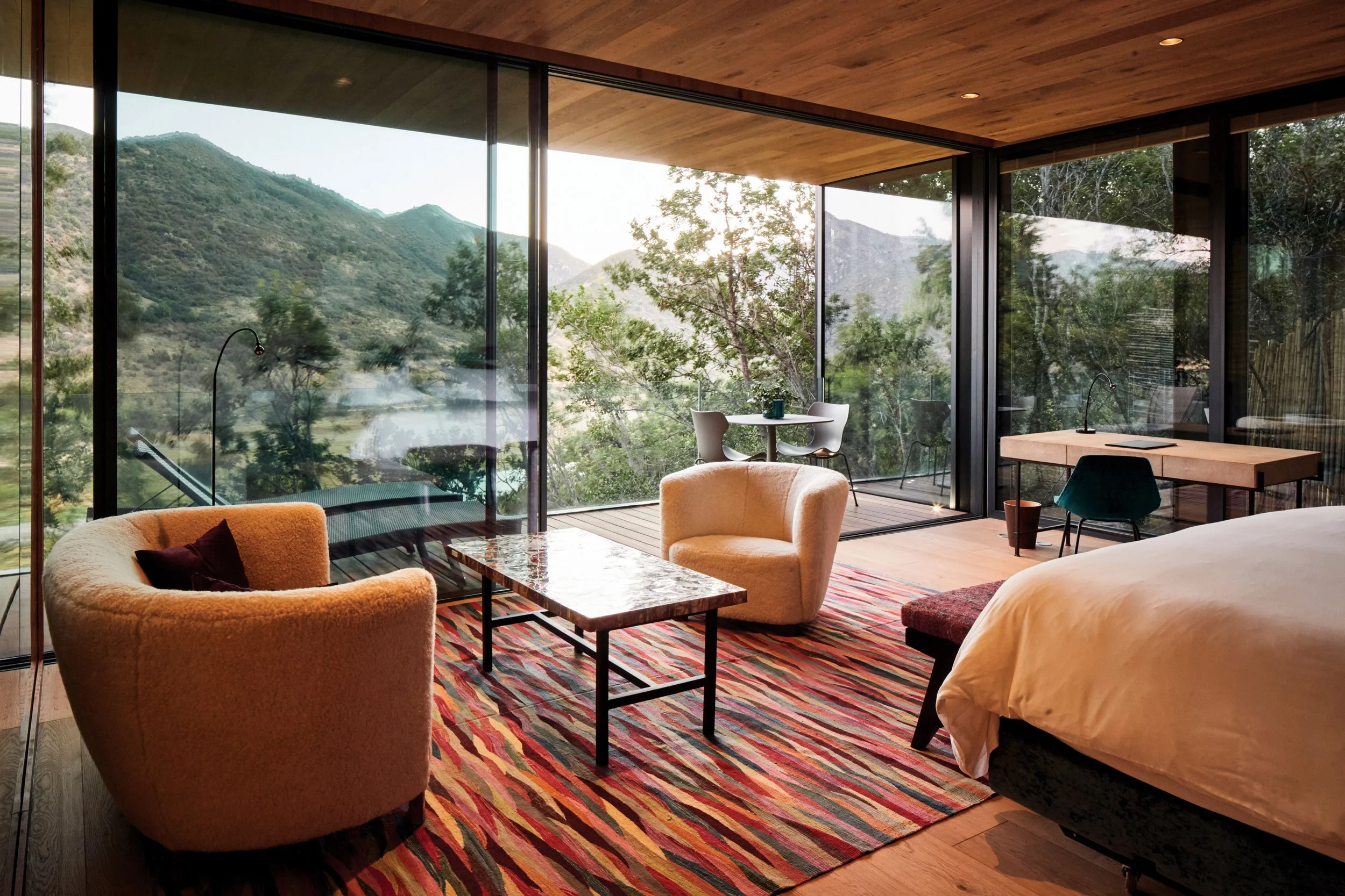 Interior de una habitación moderna con paredes de vidrio que muestran una vista de montañas y árboles, muebles de color neutro, una alfombra multicolor y madera en techos y pisos.