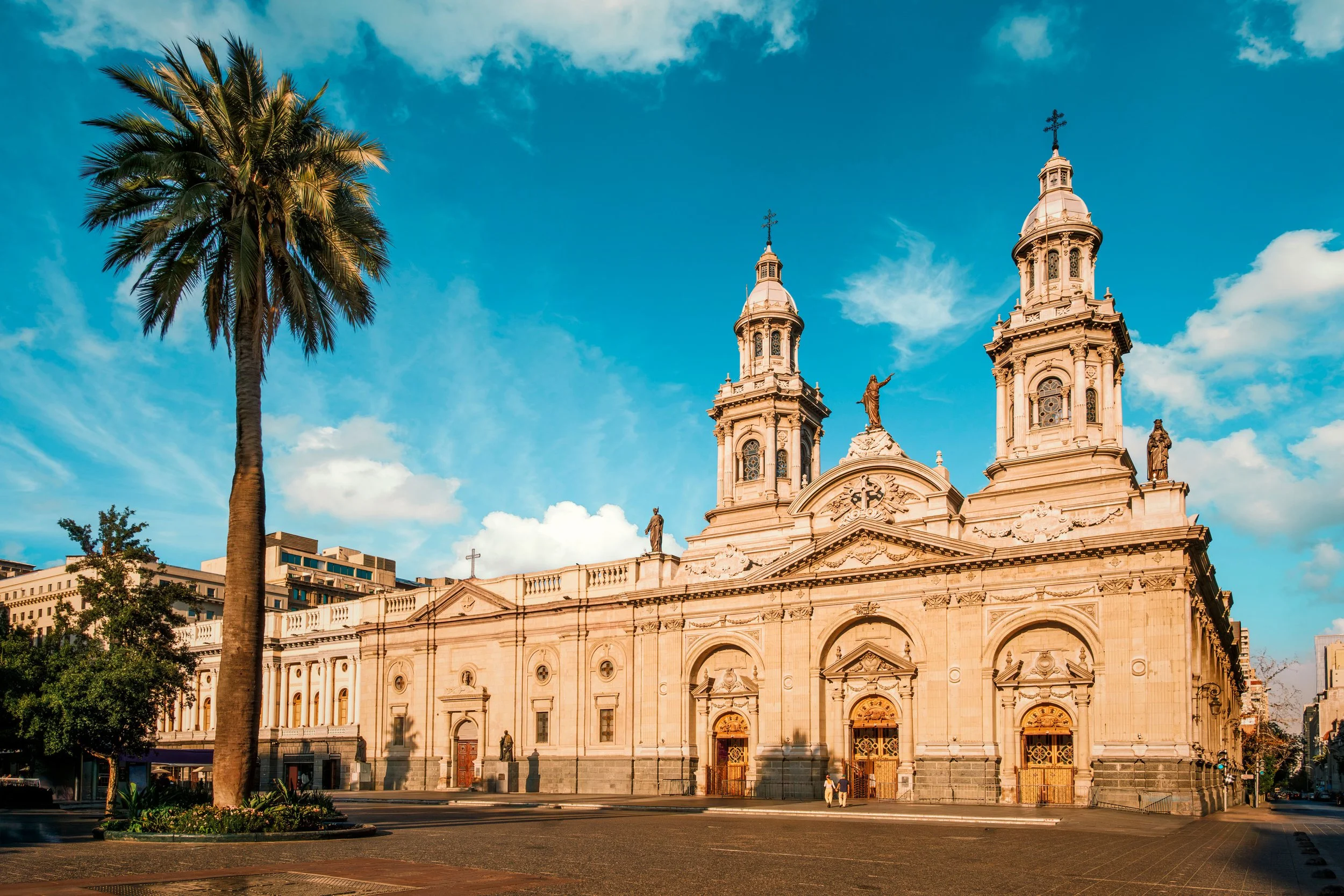 Una iglesia grande de estilo clásico con dos torres y esculturas, en una ciudad con edificios modernos circundantes, bajo un cielo despejado con algunas nubes.