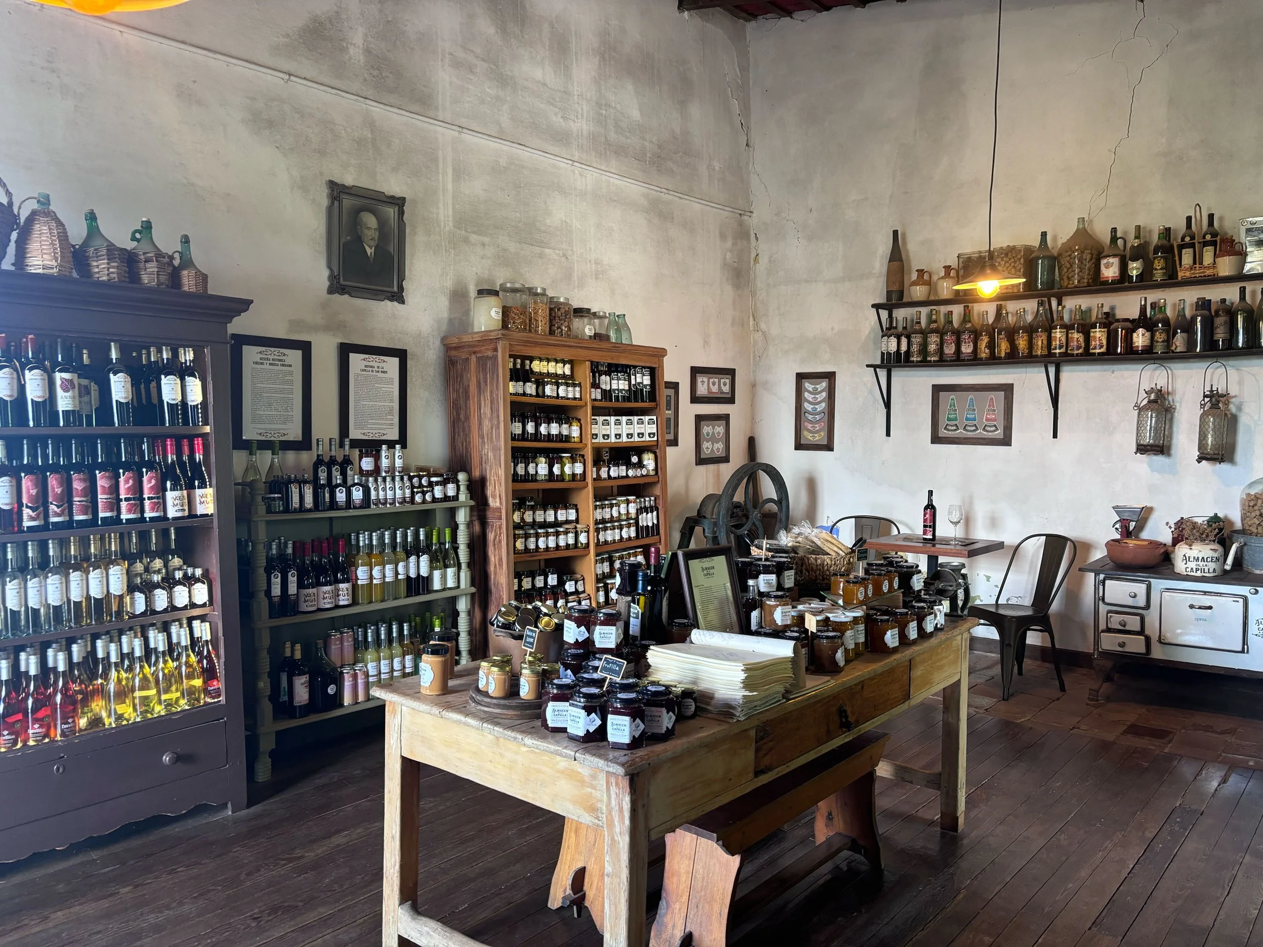 Interior de una tienda o bodega que vende productos artesanales y comestibles, con estantes llenos de frascos, botellas y otros productos, decoraciones en las paredes y una mesa de madera con algunos objetos sobre ella.