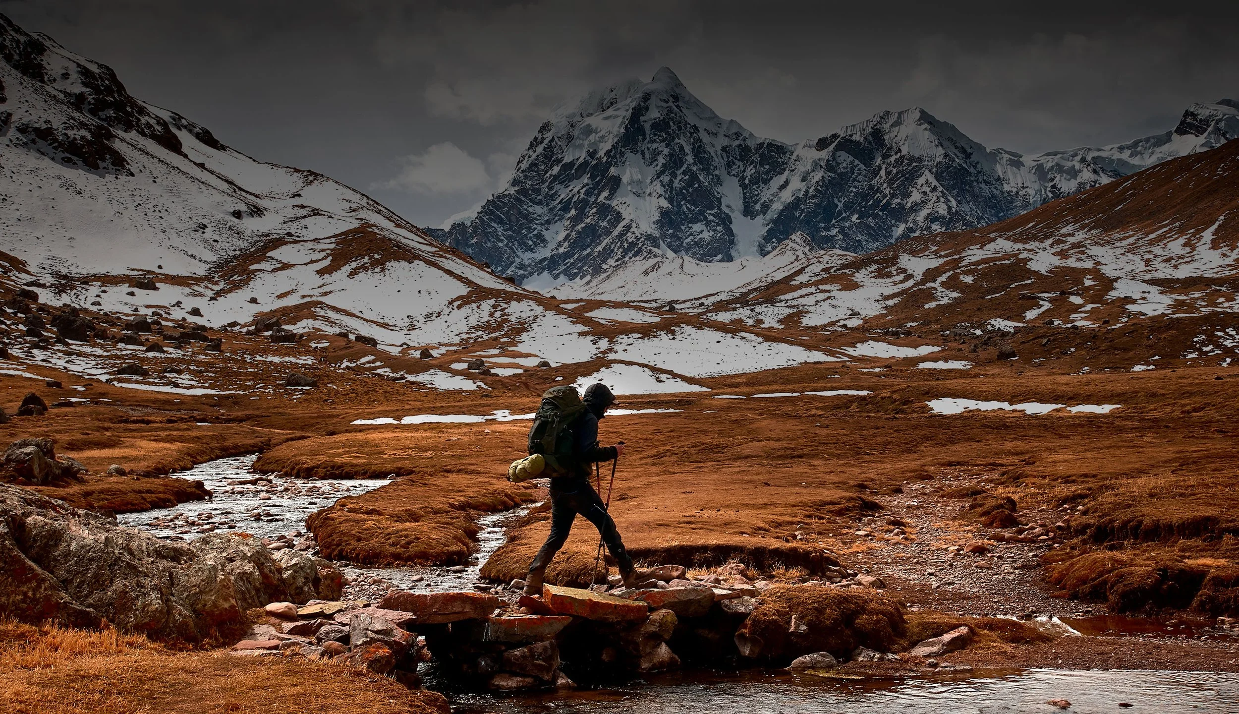 Una persona con mochila y bastón de senderismo caminando por un paisaje montañoso con nieve, árboles y un río, bajo un cielo nublado.