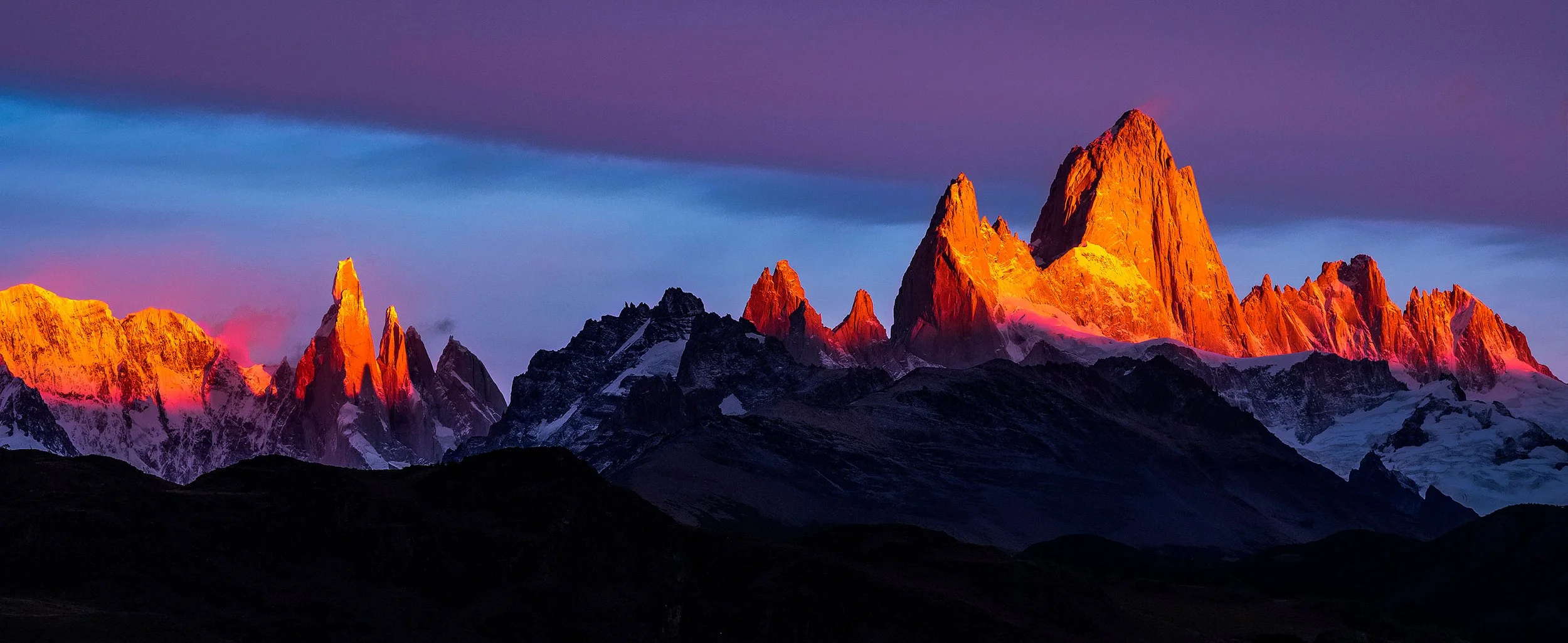 Montañas rocosas y nevadas iluminadas por el atardecer con cielo azul y nubes moradas.