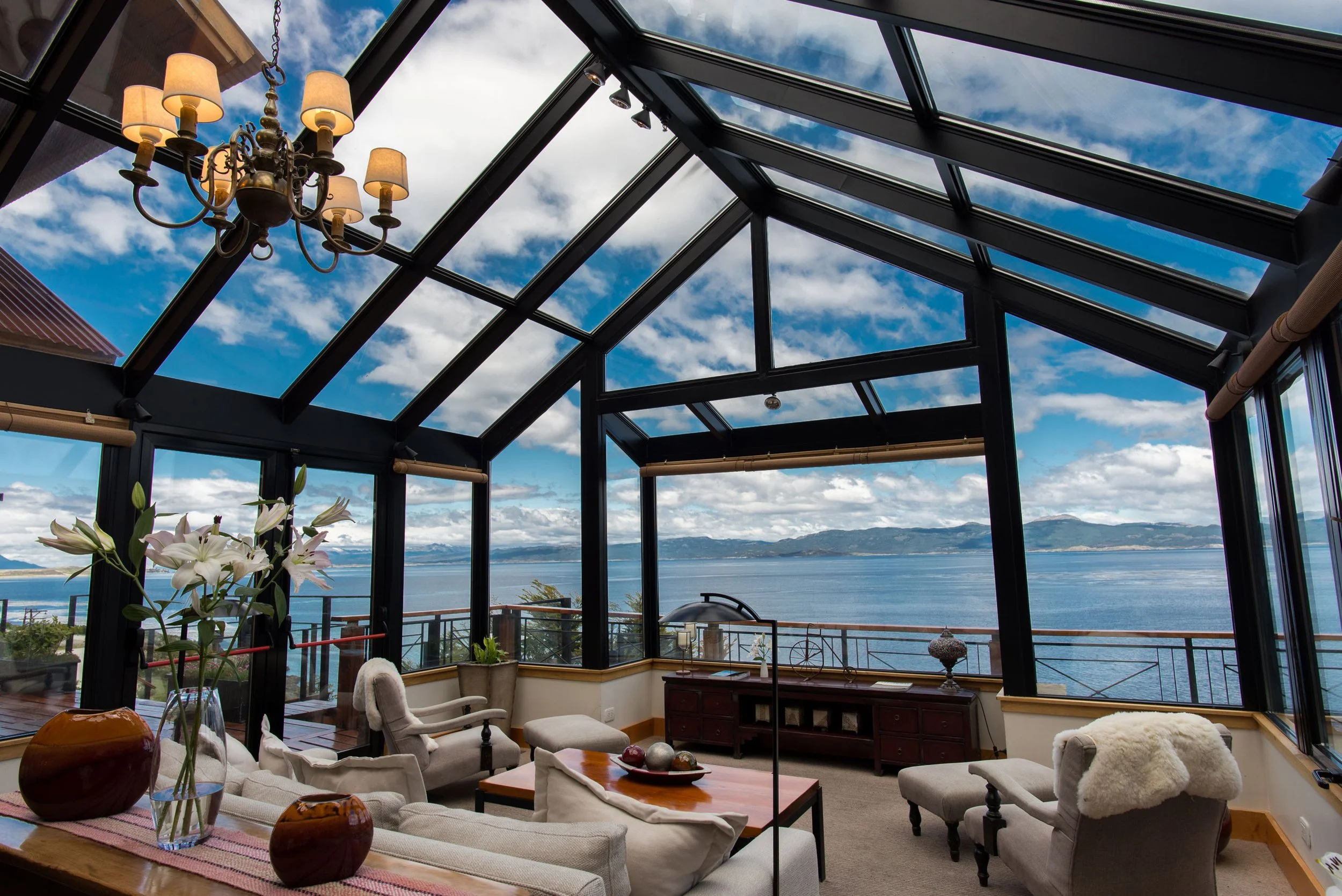 Salón con ventanas de techo de cristal, vistas al lago y las montañas, decorado con muebles blancos, una lámpara de araña, flores y objetos decorativos.
