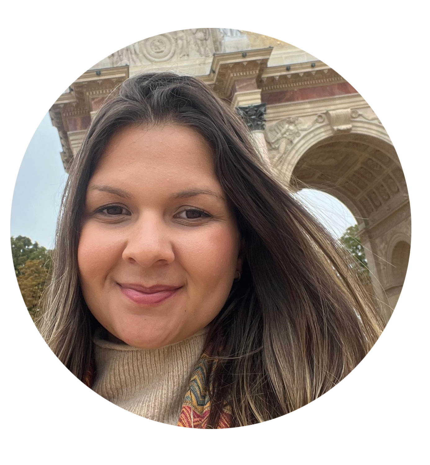 Una mujer con cabello largo y rubio claro, sonriendo, frente a un arco arquitectónico con detalles decorativos y columnas, en un entorno al aire libre.
