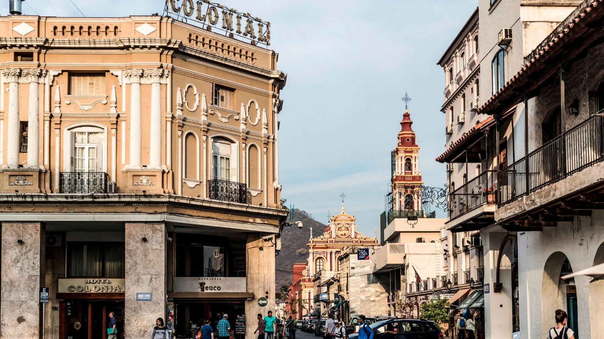 Calles de una ciudad con edificios antiguos, una iglesia en el fondo y varias personas caminando.