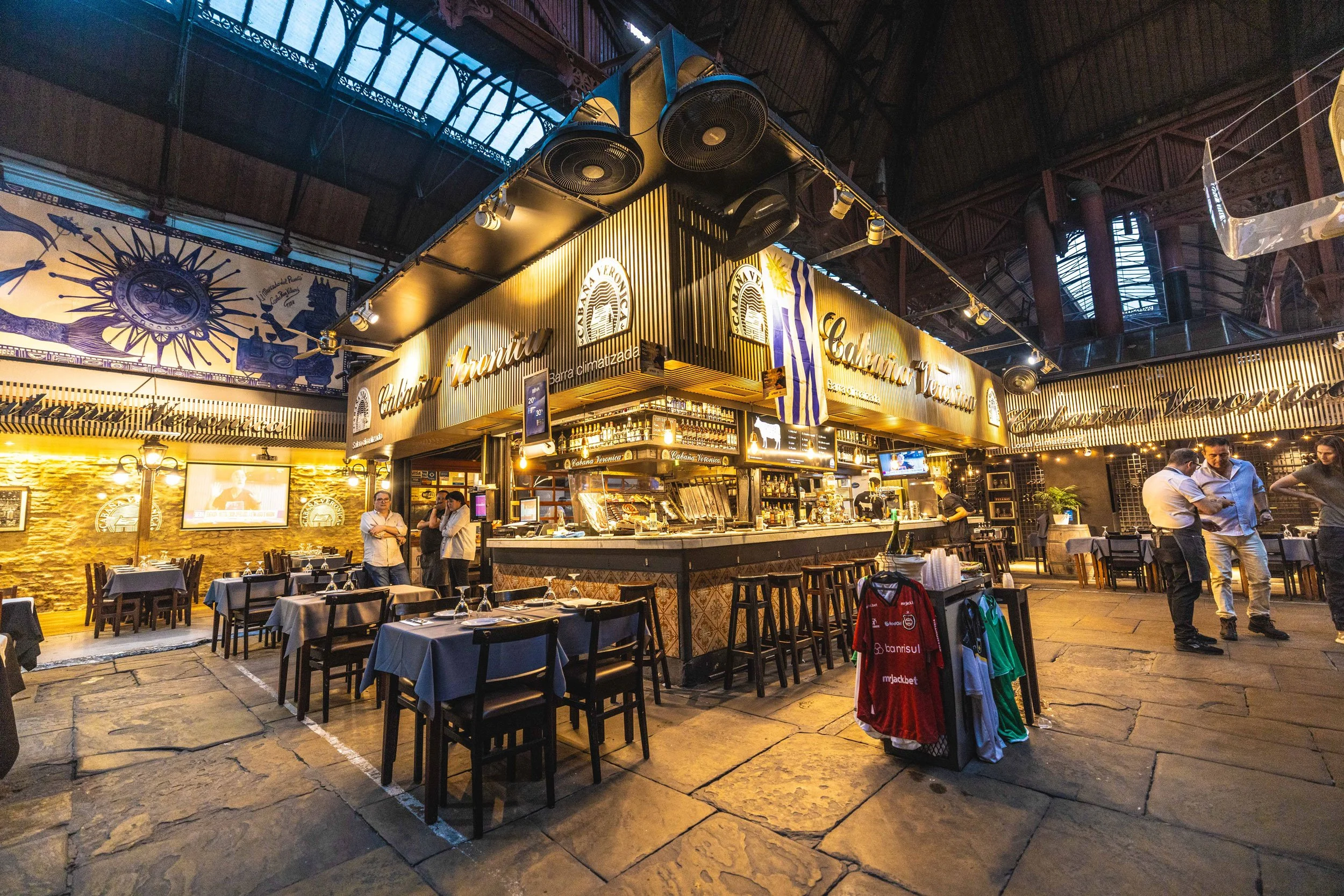 Interior de un restaurante con mesas y sillas, barra de bebidas y decoraciones en tonos cálidos, con algunas personas circulando y decoraciones con motivos mexicanos.