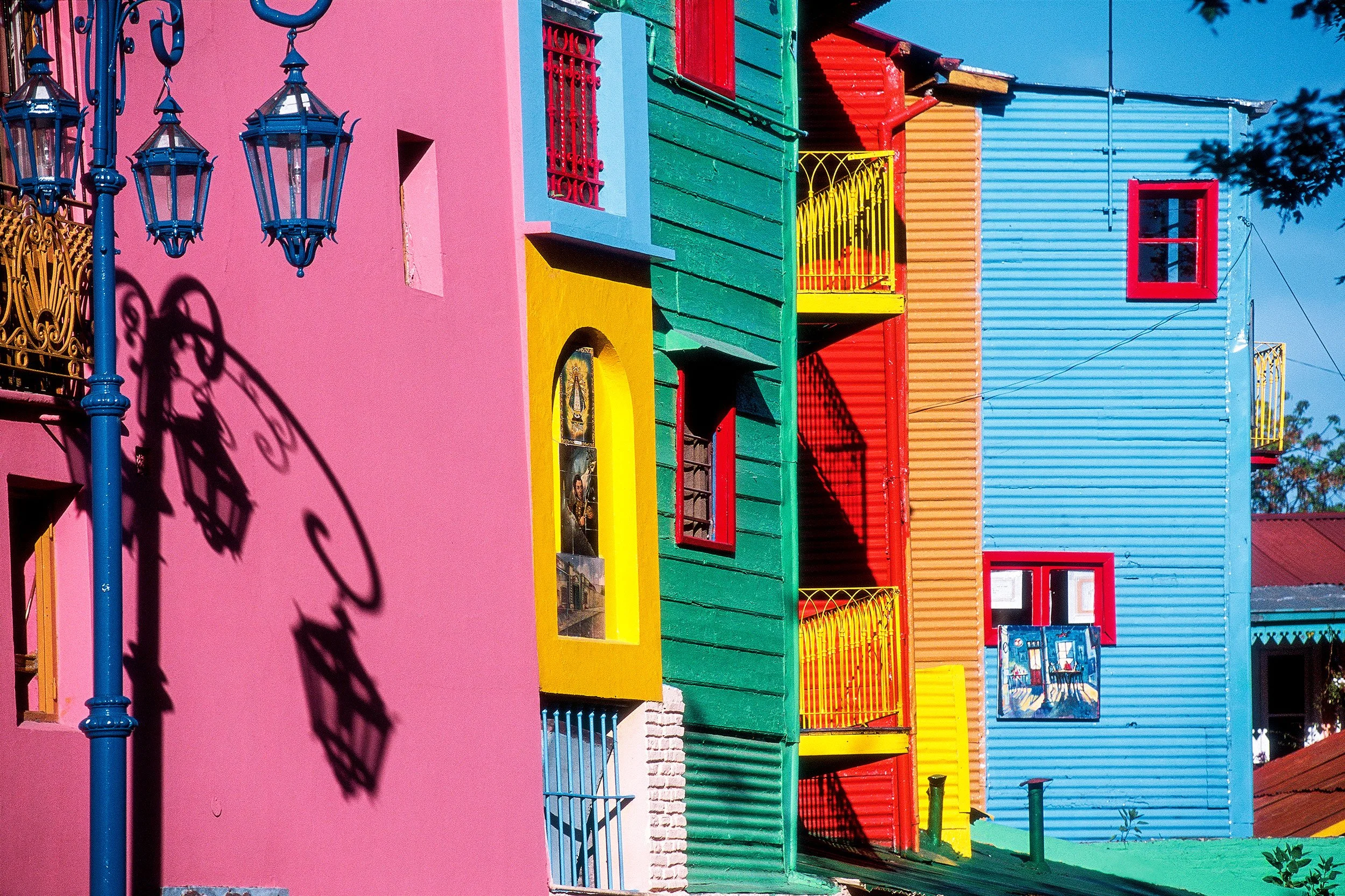 Edificio de colores en tonos rosa, azul, verde, amarillo y rojo con ventanas y balcones ornamentados, sombra de lámpara en la pared rosa, y árbol en la esquina superior derecha.