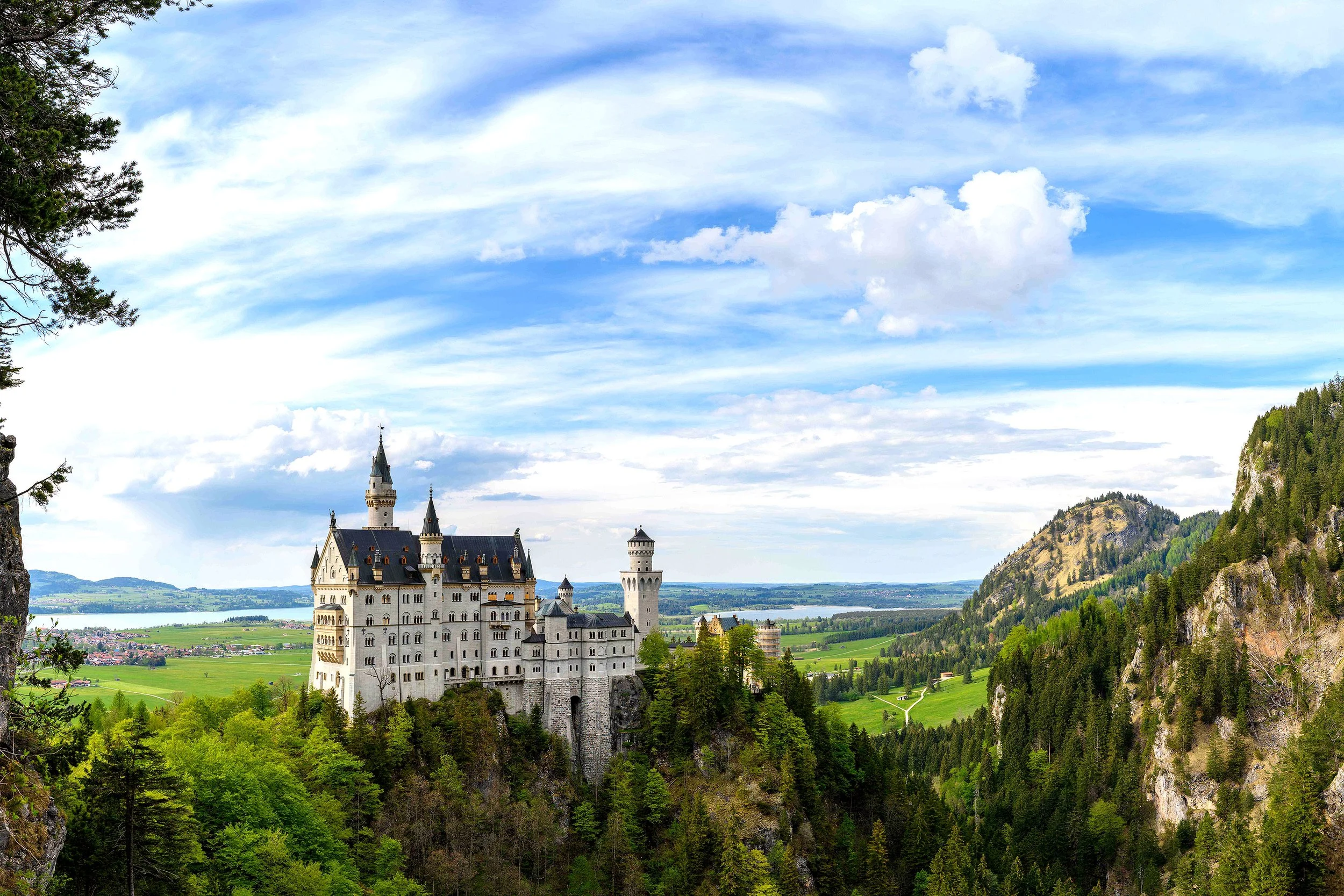 Castillo en un paisaje montañoso con bosques verdes, junto a un río y un cielo nublado con algunas nubes blancas.