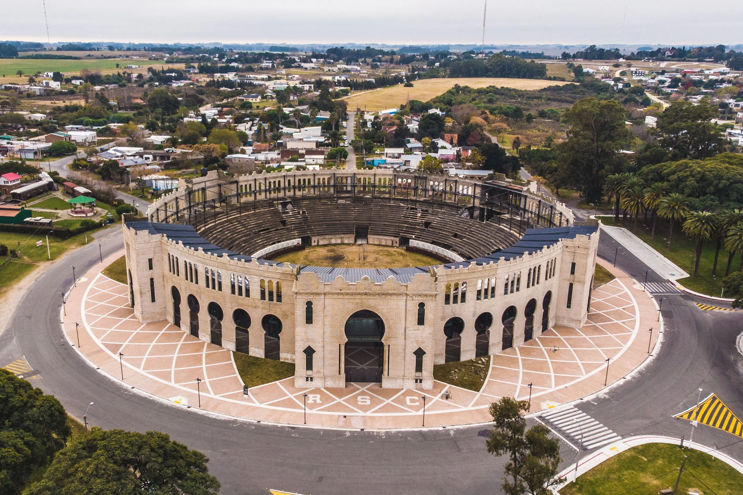 Vista aérea de un teatro antiguo con arquitectura clásica y un gran escenario, rodeado de calles y áreas verdes en un entorno suburbano.