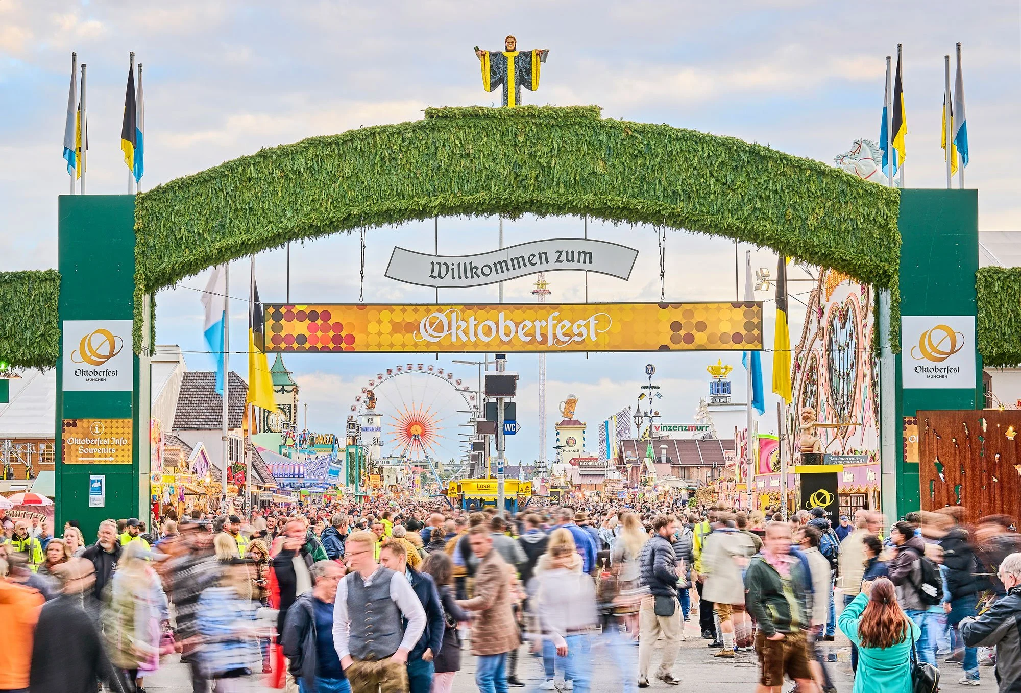 Entrada principal a la feria de Oktoberfest en Múnich, con una gran multitud de personas, un arco decorativo cubierto de vegetación, banderas de Alemania y azul y blanco, y en el fondo una noria gigante y otras atracciones de feria.