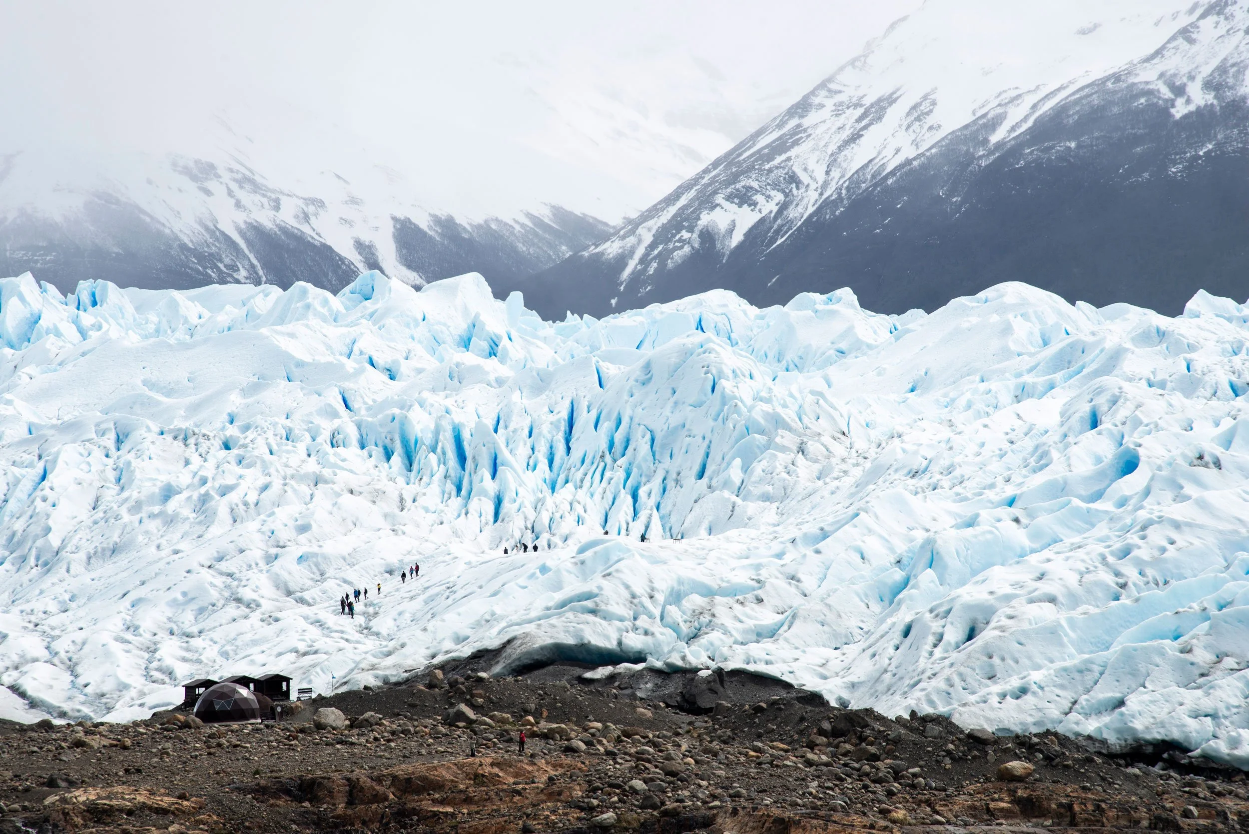 Vista de un glaciar con montañas nevadas al fondo y un grupo de personas caminando sobre hielo.