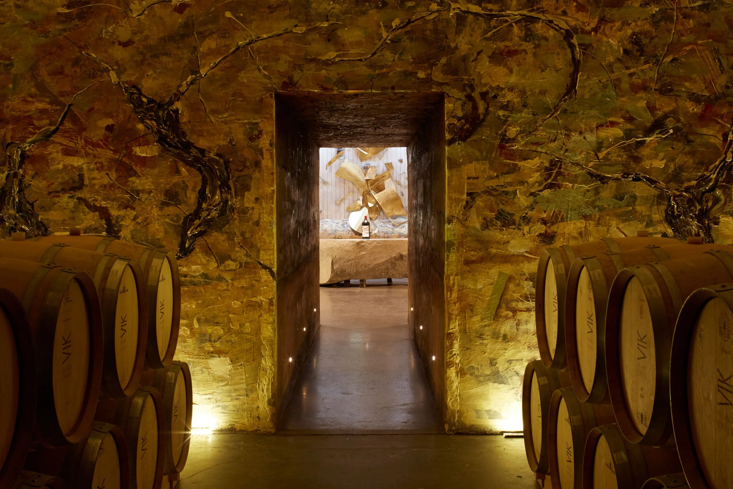 Imagen de una bodega de vino con barriles de madera alineados a los lados, una entrada en el fondo con paredes decoradas con trozos de madera y un vaso de vino con botella en el centro.