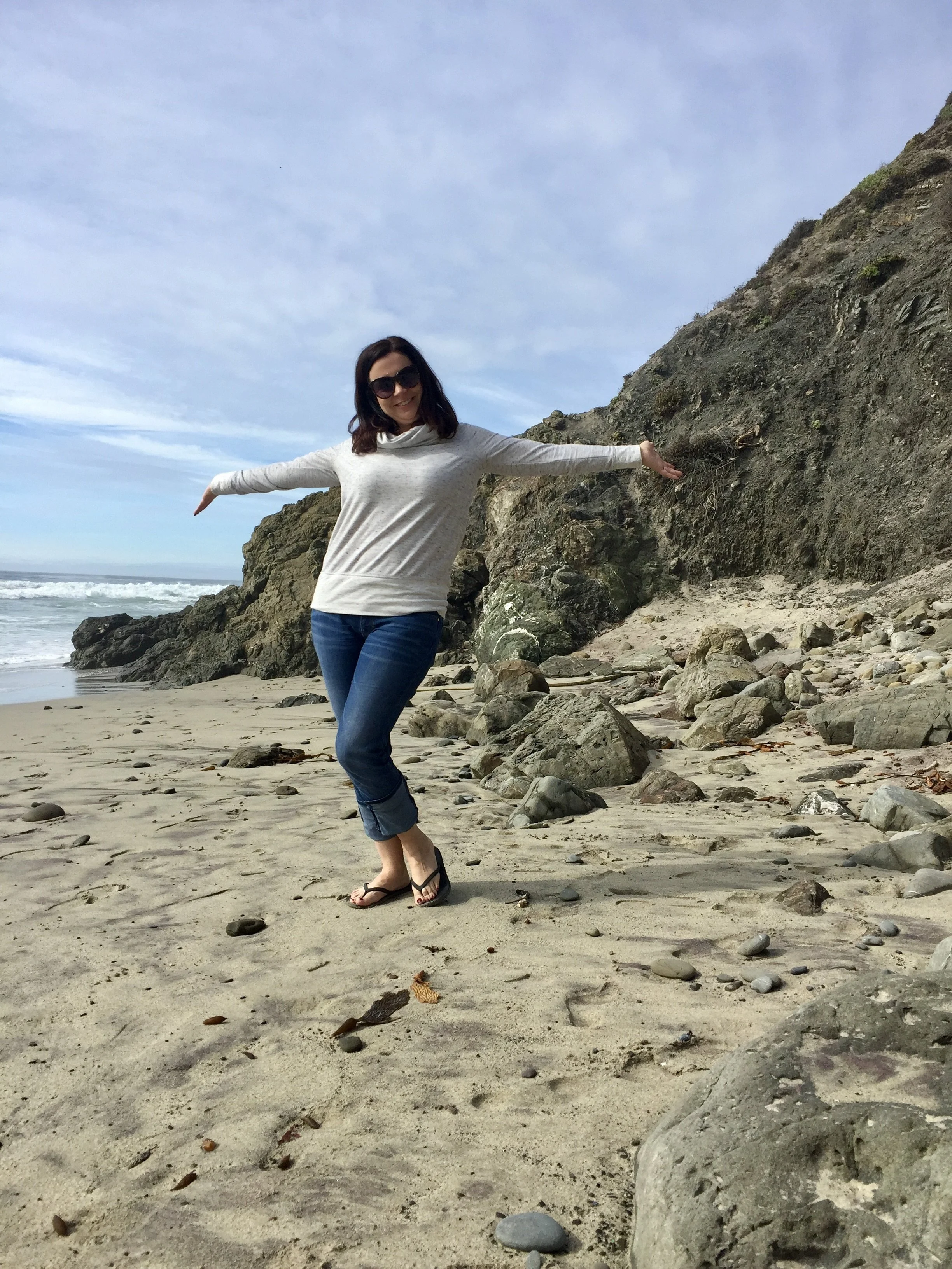 Jodi Barnum posing with outstretched arms on a rocky beach with ocean waves and a cloudy sky.
