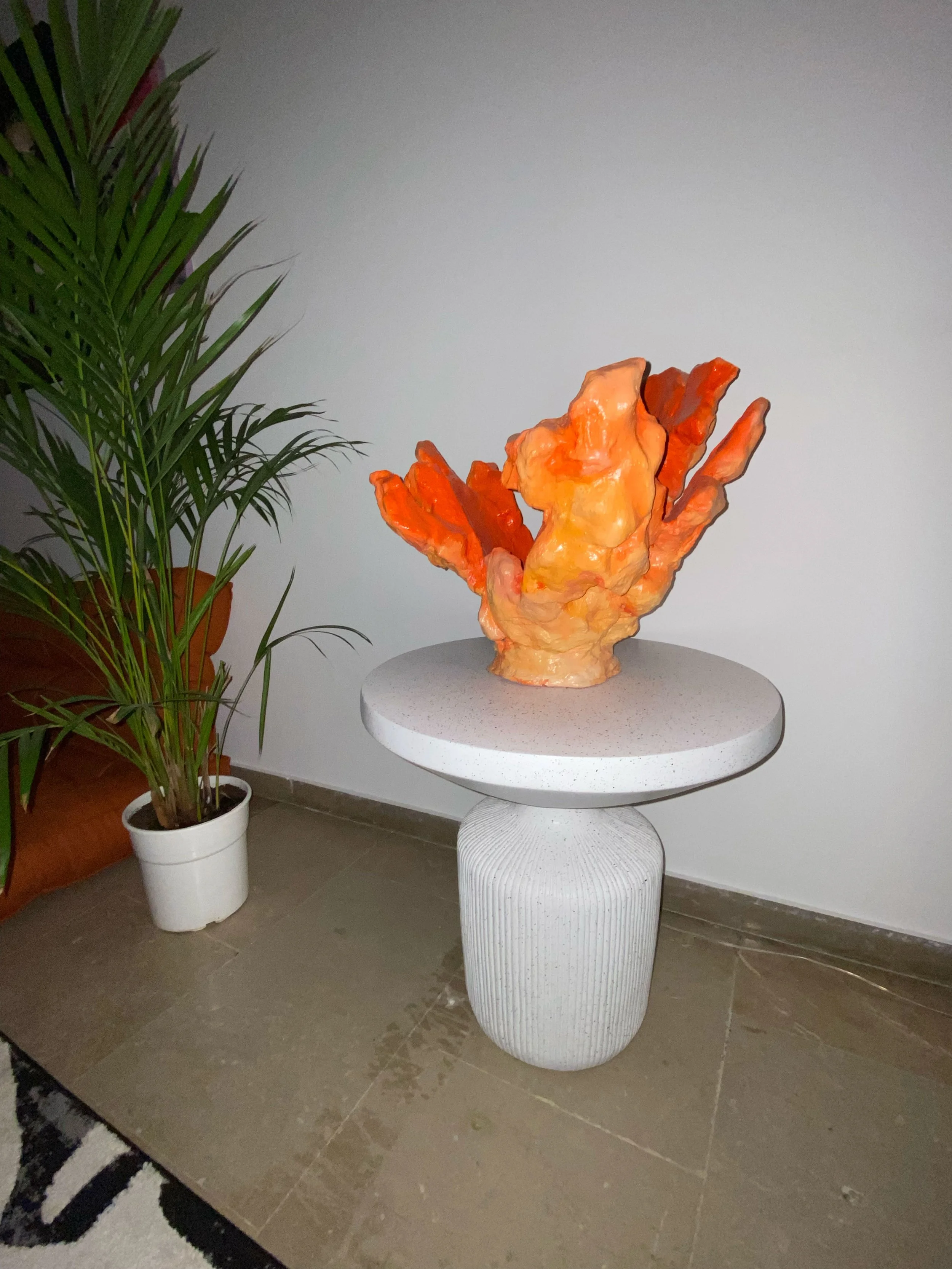 A modern decorative sculpture with orange and yellow hues on a round white table with a textured cylindrical base. A potted green plant with long, slender leaves is positioned on the floor to the left of the table. The scene is indoors with tiled flo