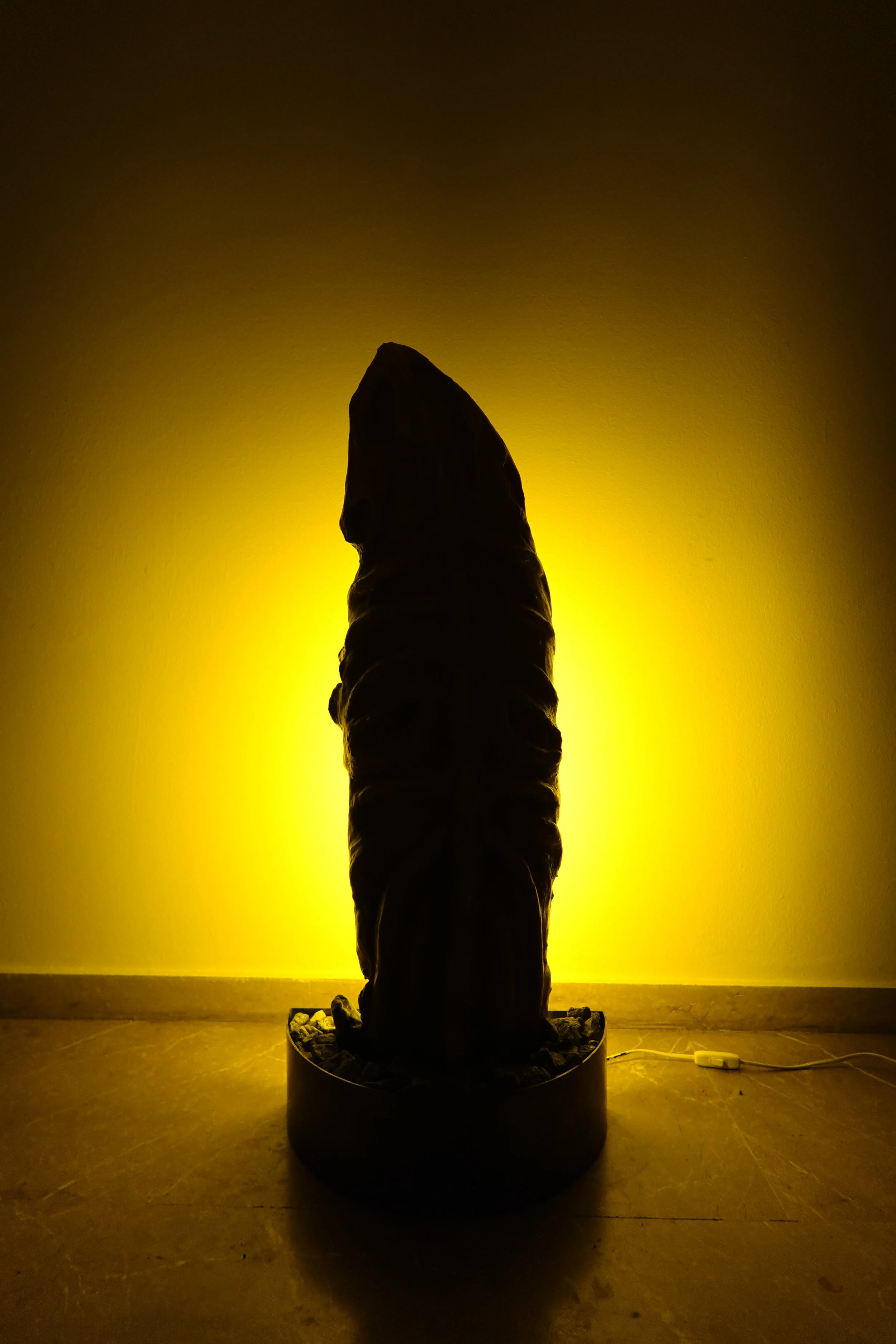 A dark sculpture of a horse's head illuminated from behind with yellow lighting, standing on a base on a wooden floor against a yellow wall.