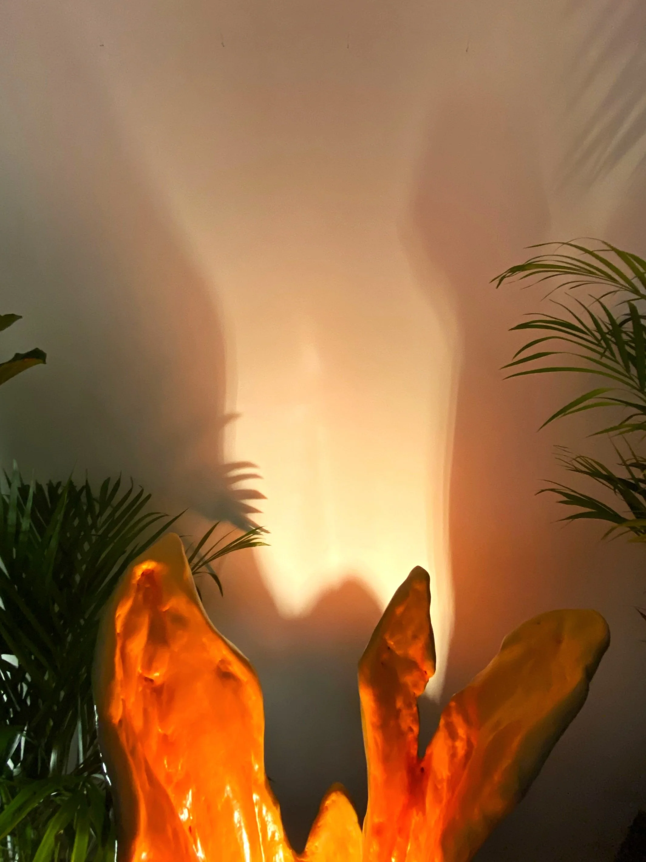 Decorative orange sculpture with a glowing, translucent appearance surrounded by green plants and casting shadows on the wall.