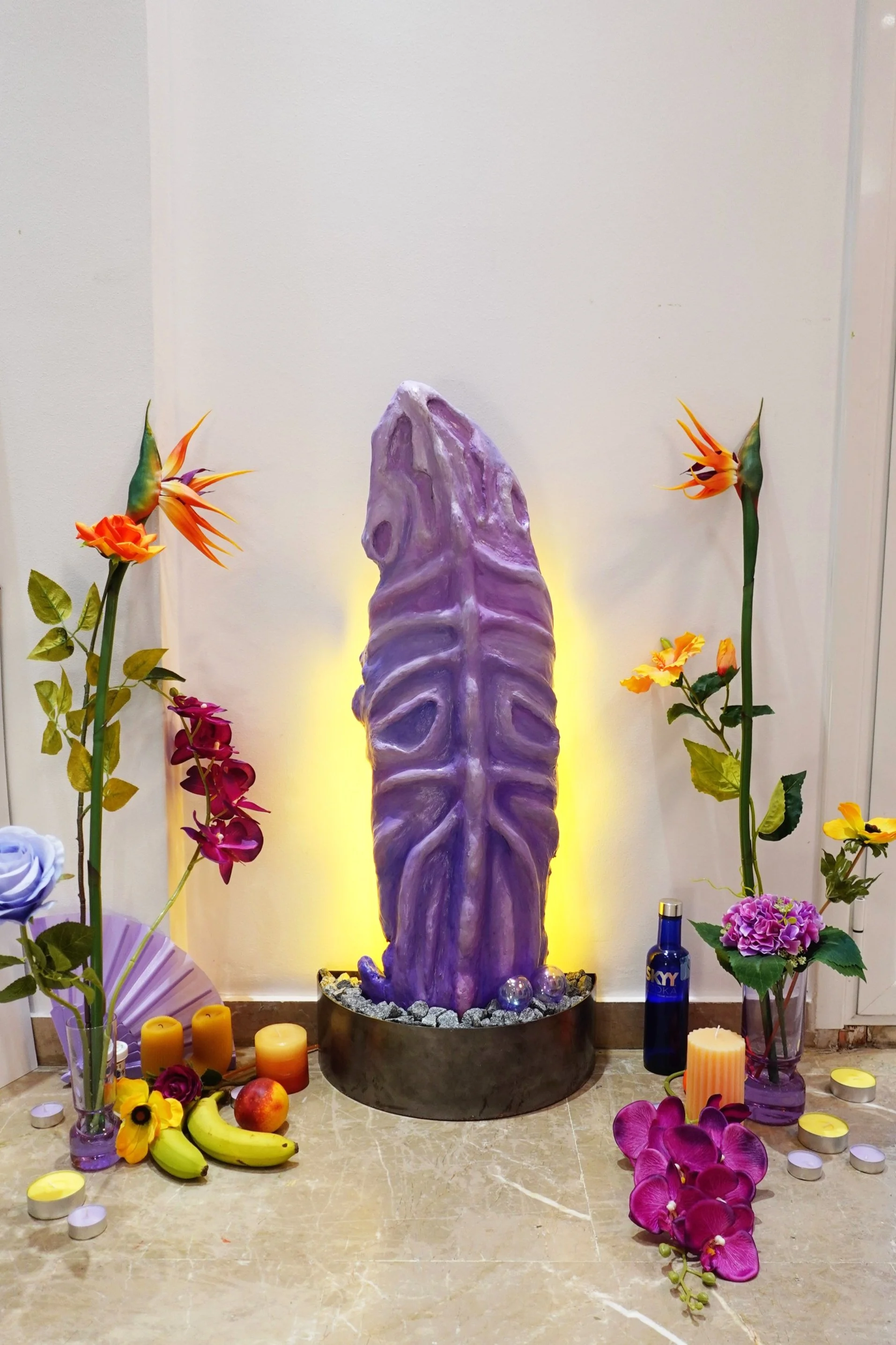 Decorative altar with a tall purple and white sculpture at the center, surrounded by colorful flowers, candles, and fruits.