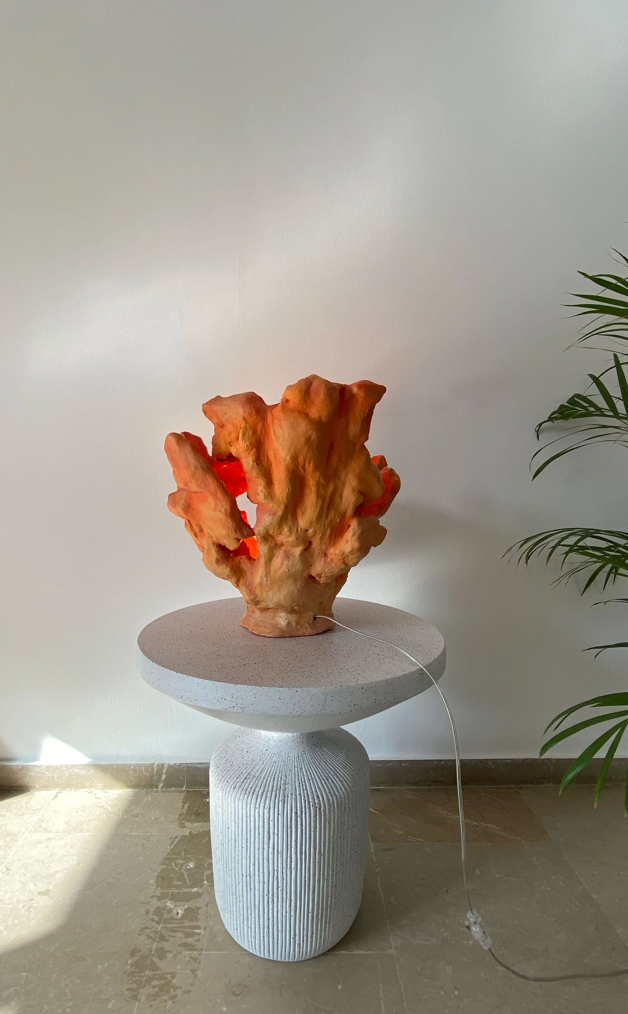 An abstract sculpture resembling a coral on a textured pedestal, placed on a round table with a cord attached, in an indoor setting with a potted plant visible on the right.
