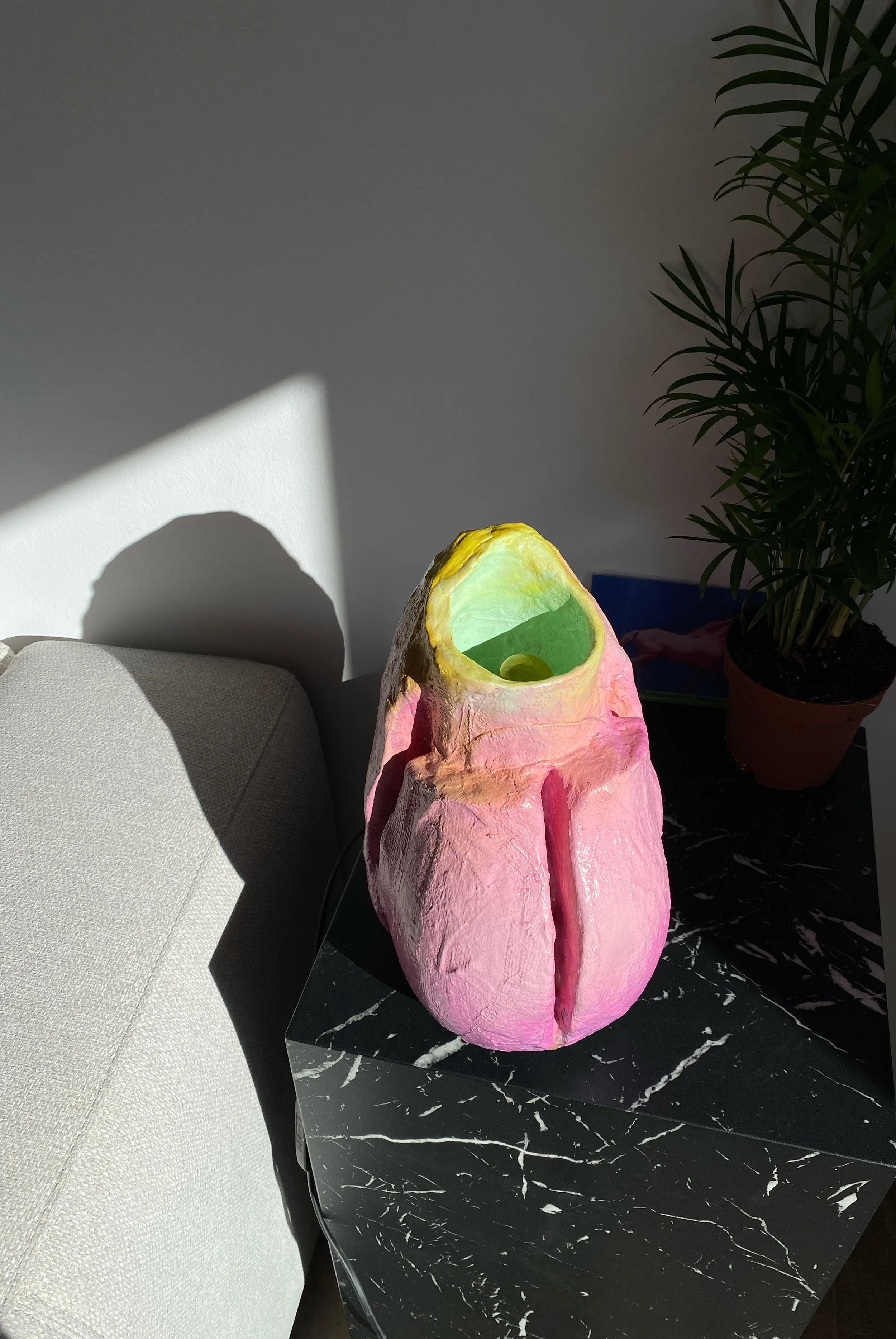 Colorful sculptural lamp on a black marble table, with a potted plant and a gray cushion nearby, illuminated by sunlight.