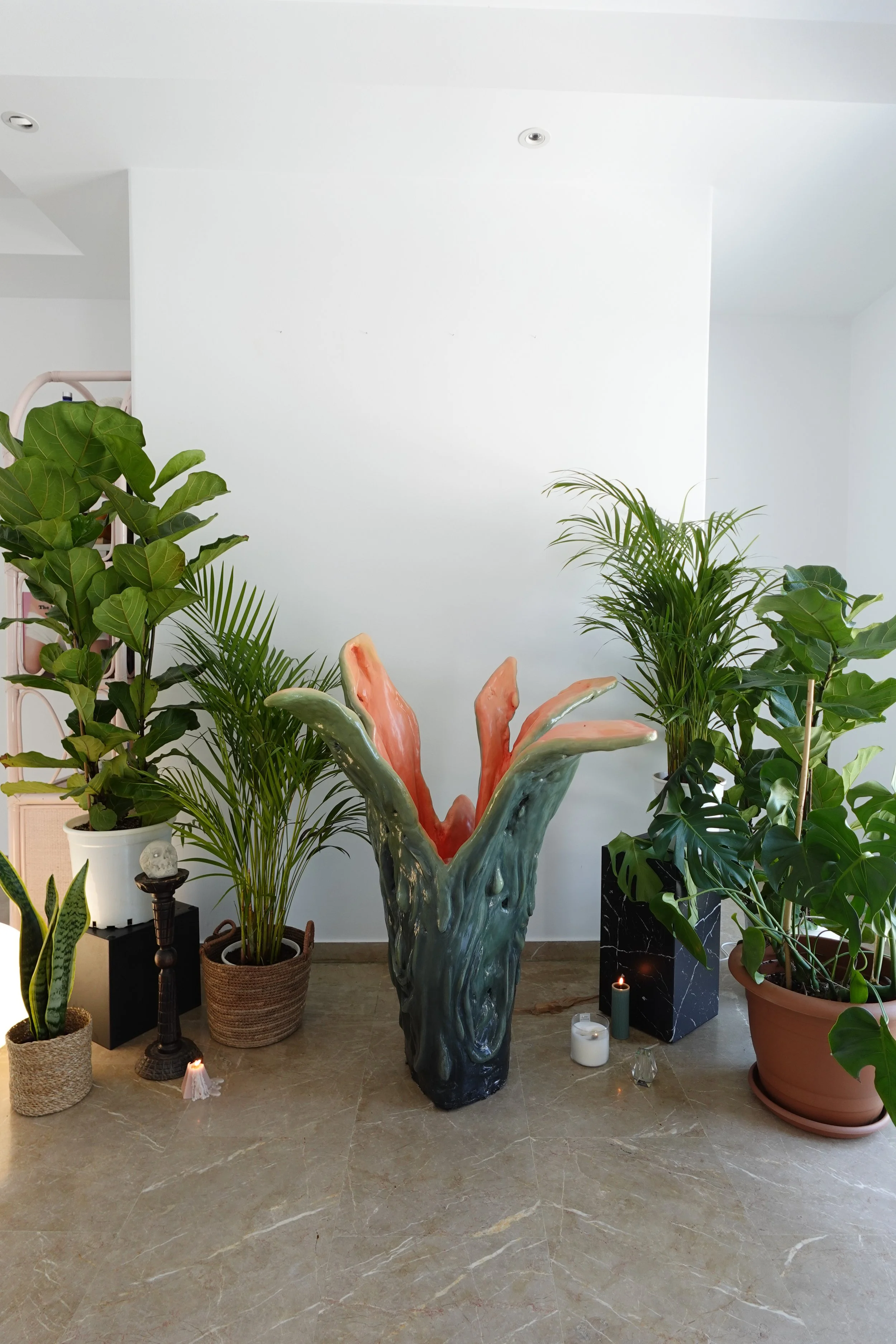 An interior scene with various green plants, candles, and a large abstract sculpture in the center. The background features a white wall.