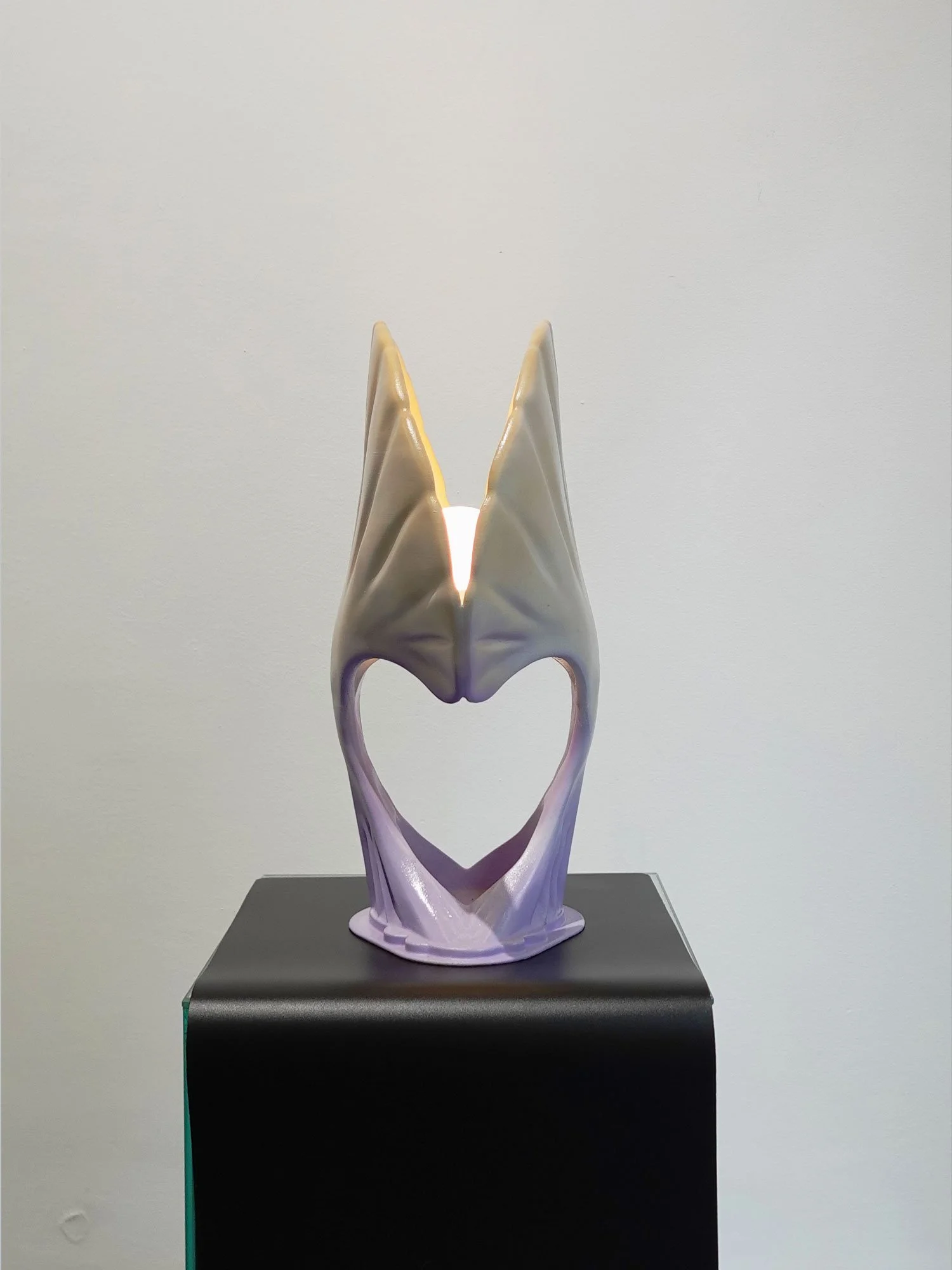 A decorative sculpture resembling a heart with wings, with lighting behind it, on a black pedestal against a plain background.