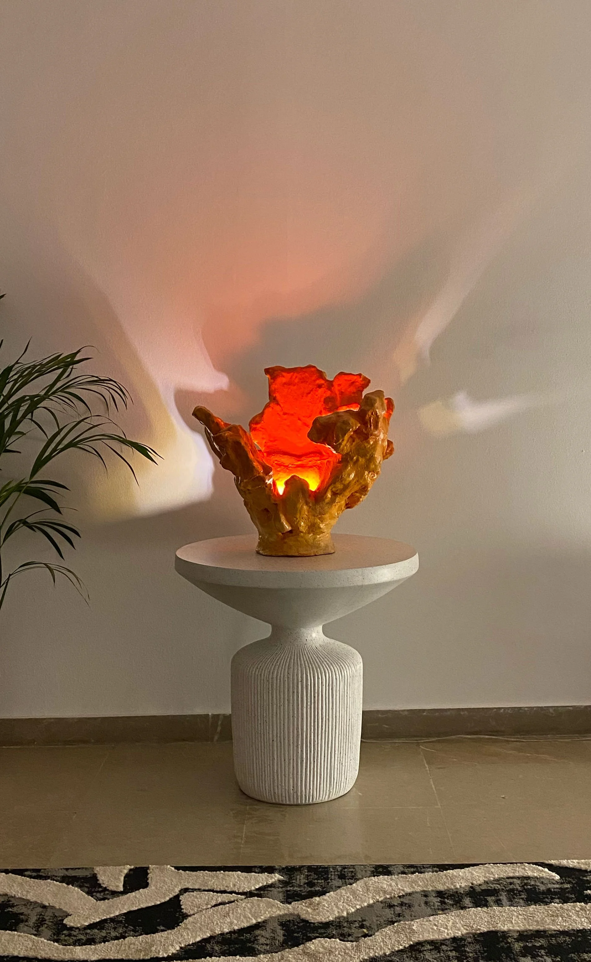 Decorative orange ceramic sculpture on a white pedestal table in a room with a plant to the left and a rug with black and white stripes.