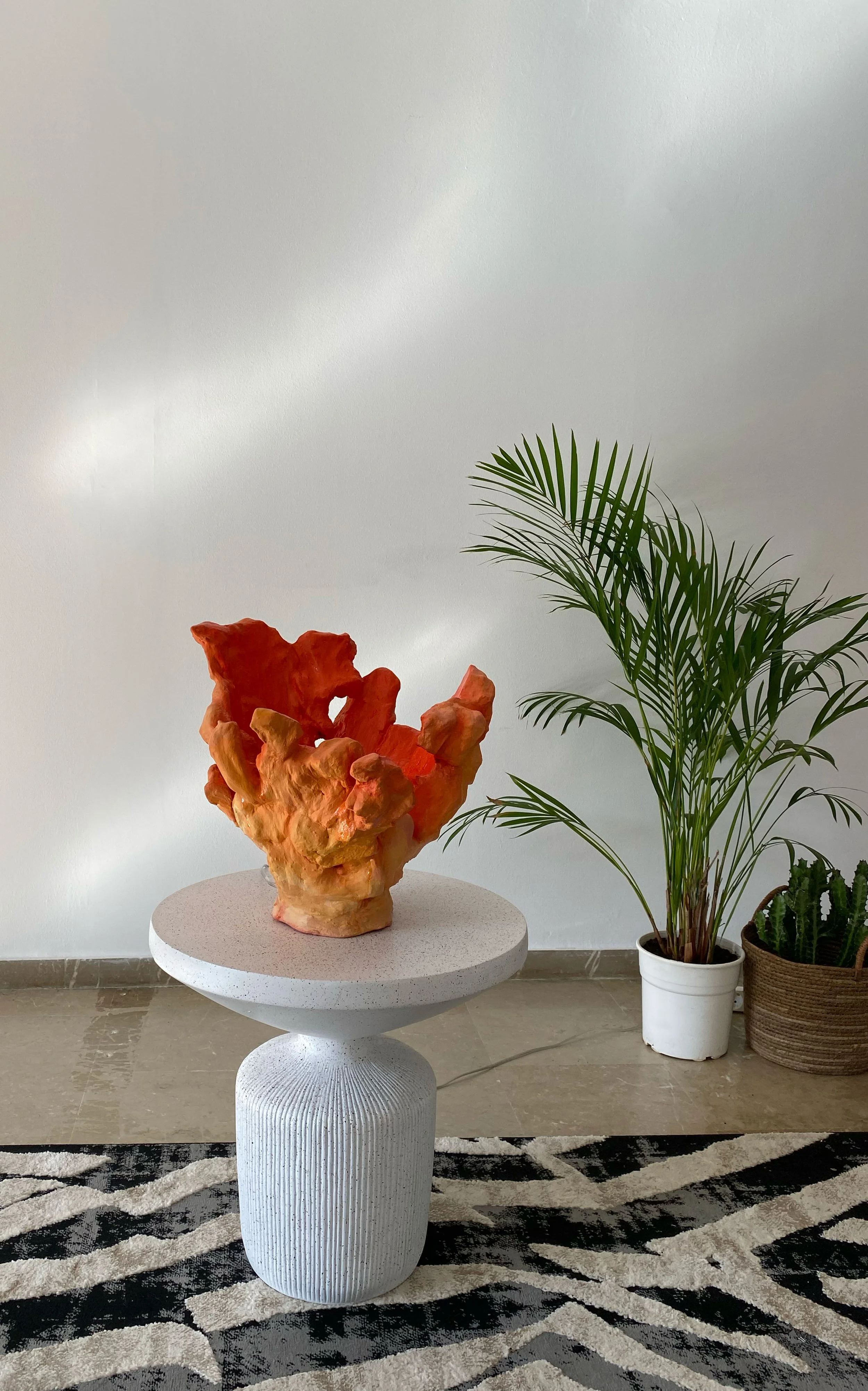 Decorative reddish-orange sculptural art piece on a white textured side table with plants in pots beside it