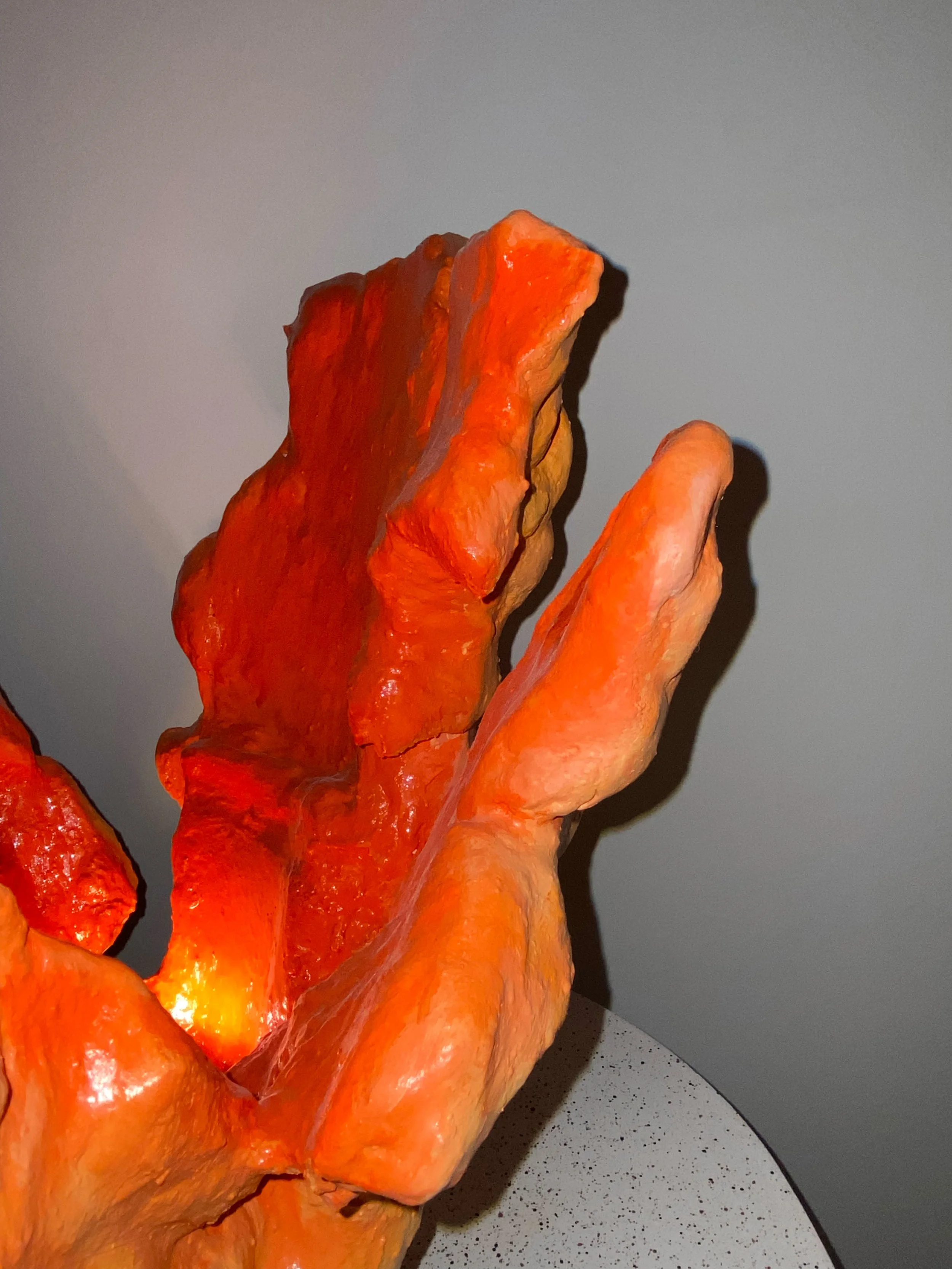 Close-up of an orange abstract sculpture resembling a hand, placed on a speckled surface with a plain wall in the background.