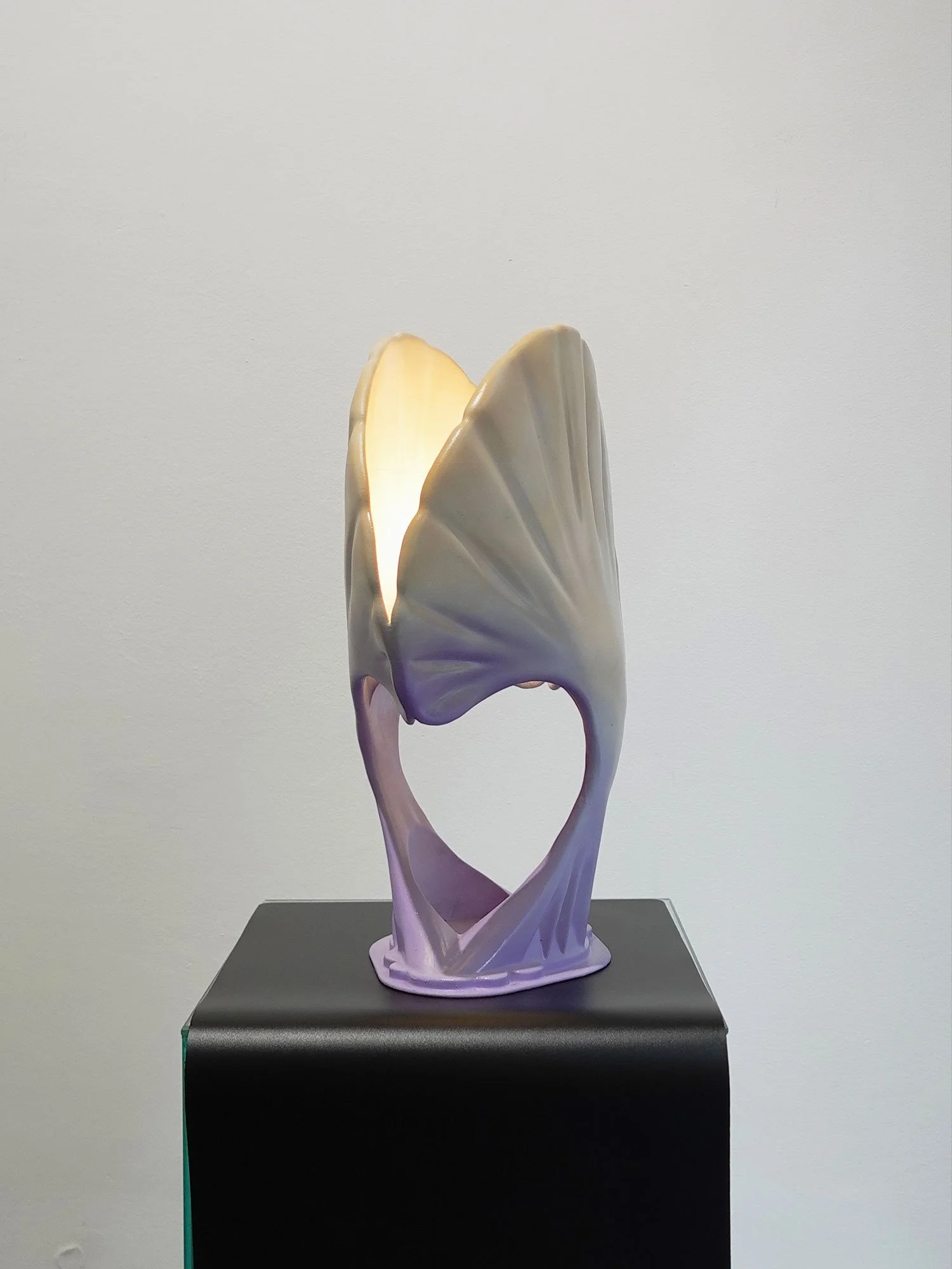 A decorative lamp with an abstract, sculptural design resembling a seashell, placed on a black pedestal against a plain wall.