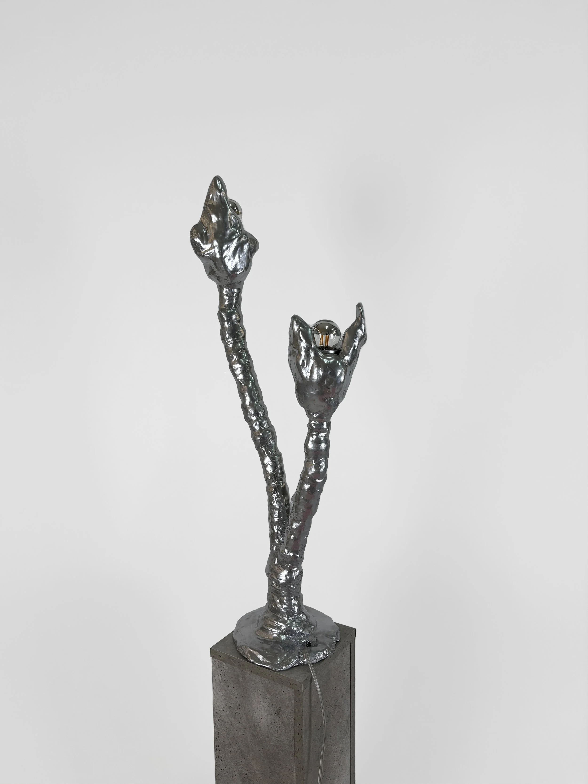 Sculpture of two metallic branches with abstract flower buds, mounted on a gray pedestal against a plain white background.