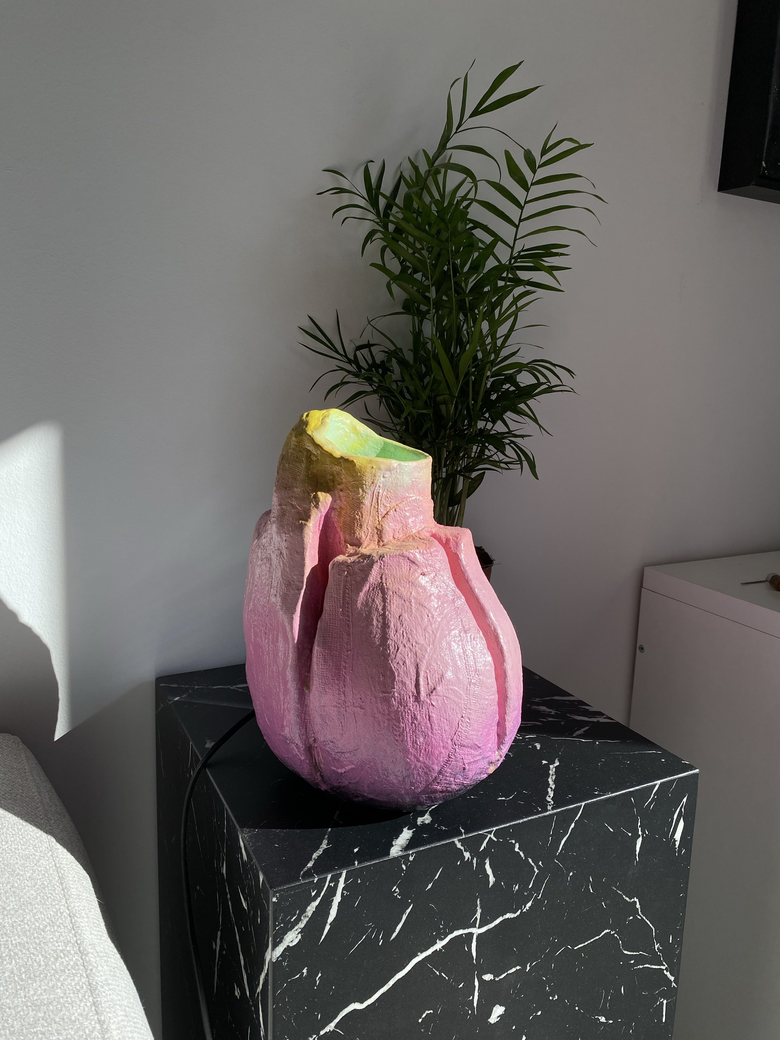 Decorative pink and yellow ceramic vase on a black marble table, with a green potted plant in the background.