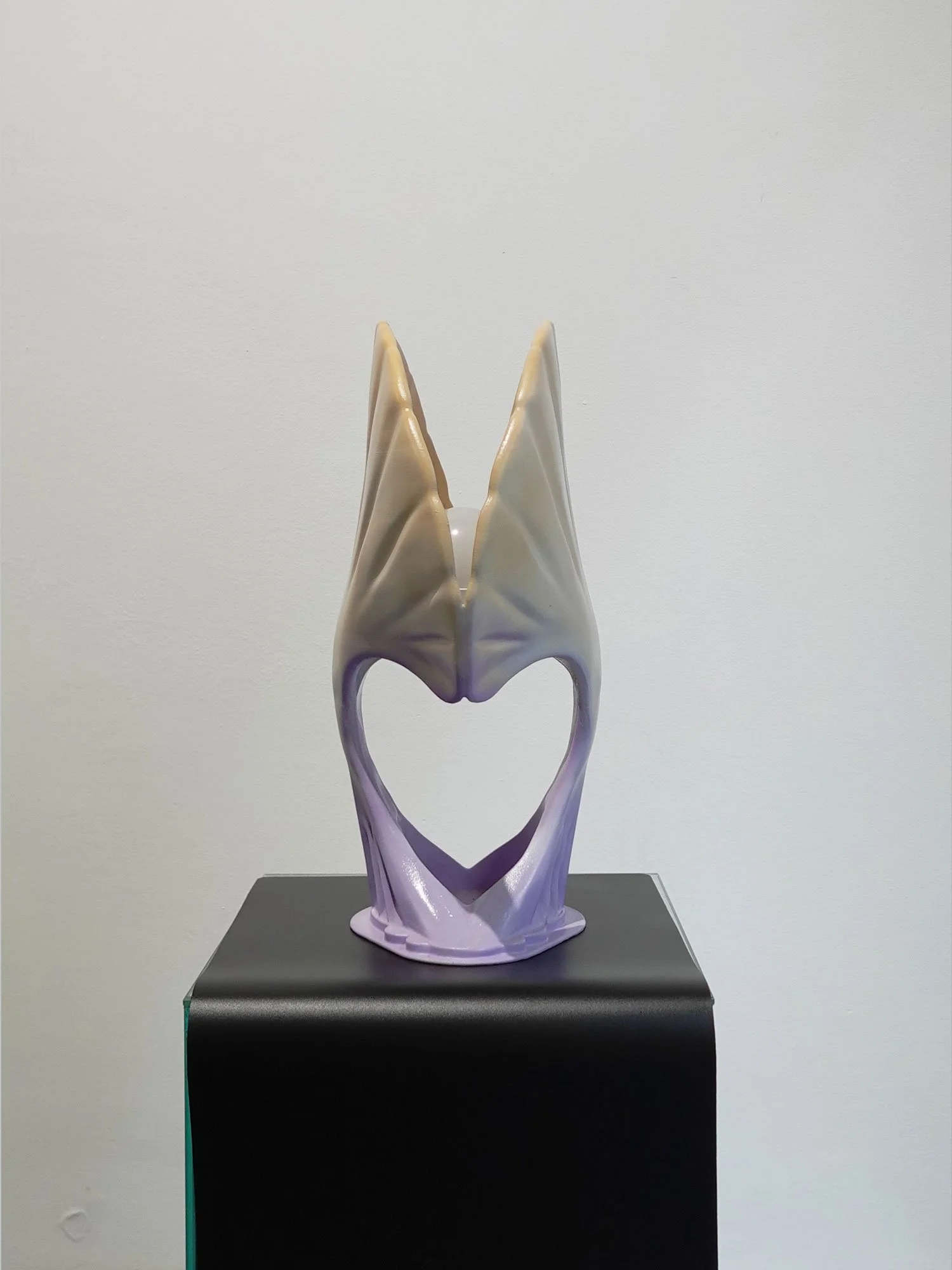 An abstract ceramic sculpture with a heart-shaped opening, featuring two elongated, pointed cream-colored wings or horns, glazed in shades of white, cream, and lavender, displayed on a black pedestal.
