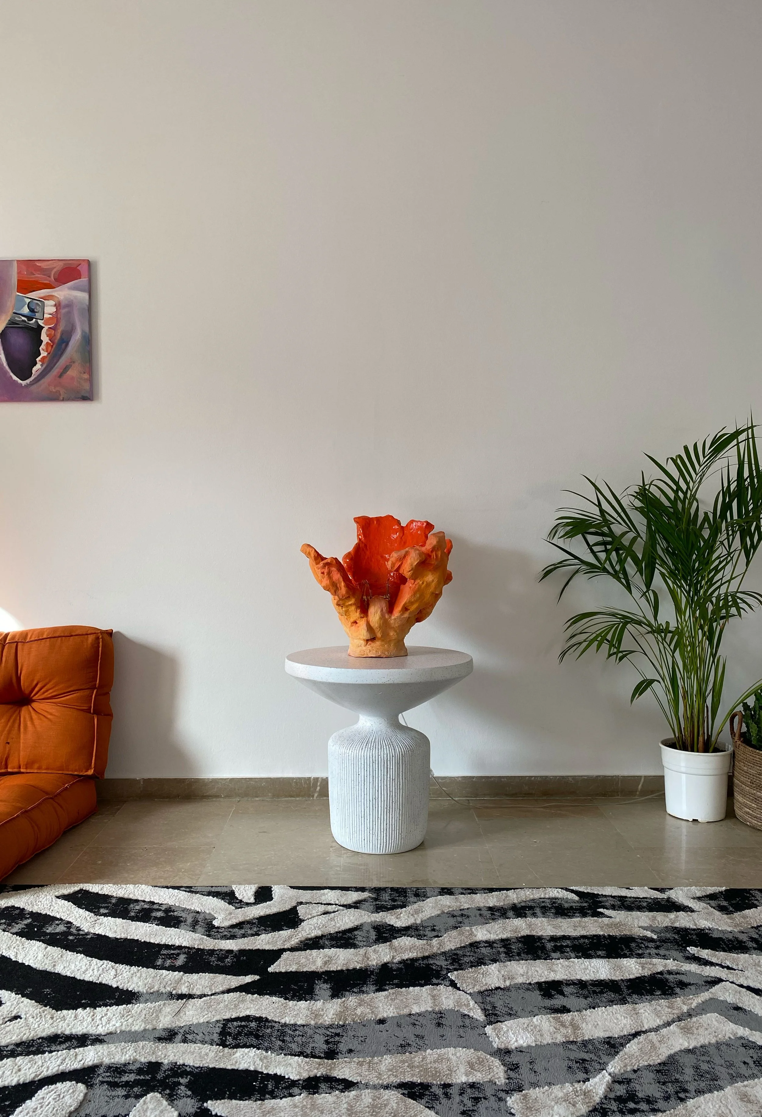 Minimalist room with a sofa, potted plant, and modern art on the wall. Central feature is a sculptural orange-red ceramic piece on a white pedestal table. Rug with black and white abstract pattern.