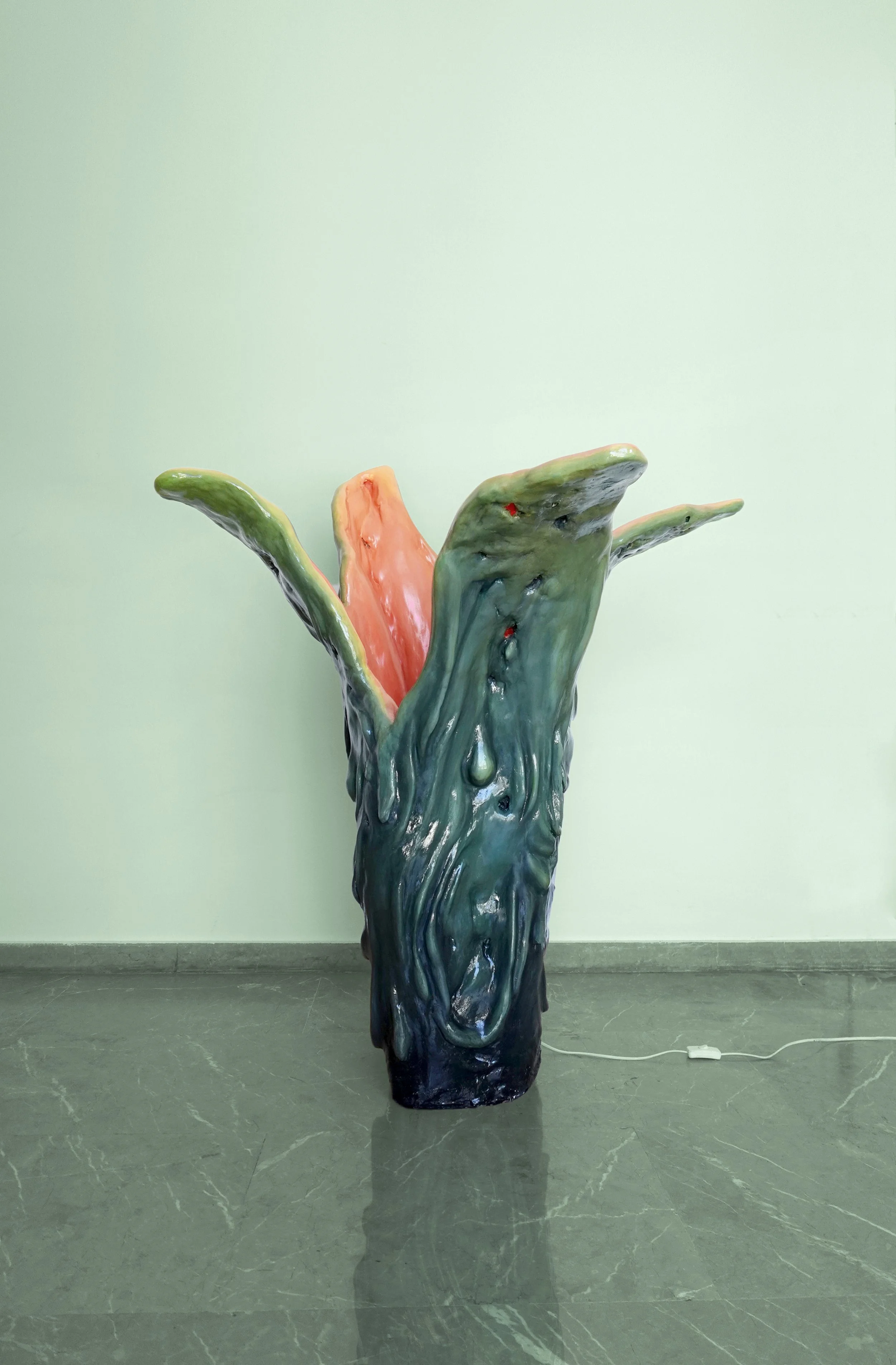Sculpture resembling a melting plant or organic form with drippy textures, green outer surface, and pink interior, standing indoors on a gray tiled floor against a light green wall.