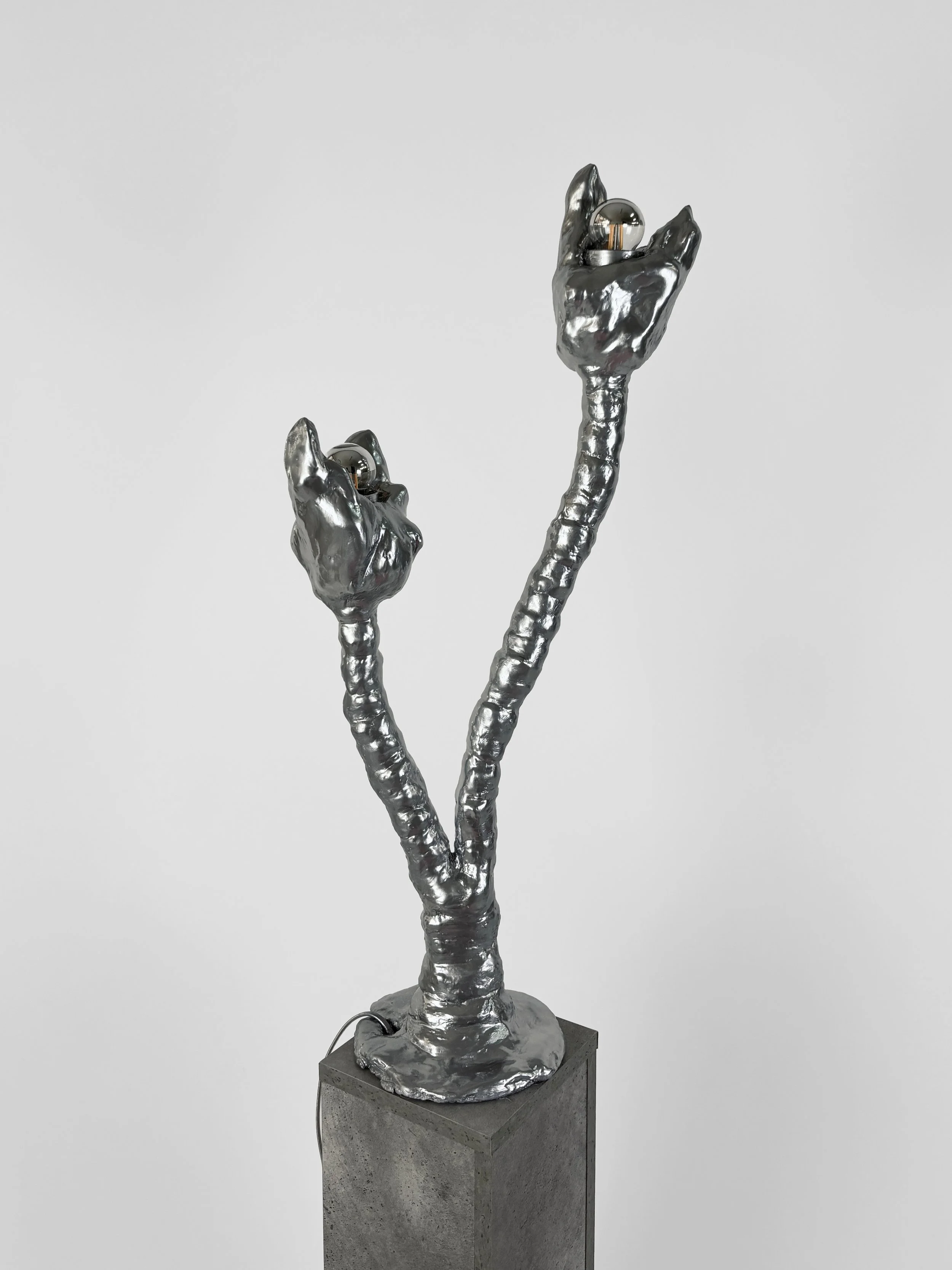 Sculpture of a metallic, silver-colored plant with two stems and flower buds, mounted on a concrete pedestal.