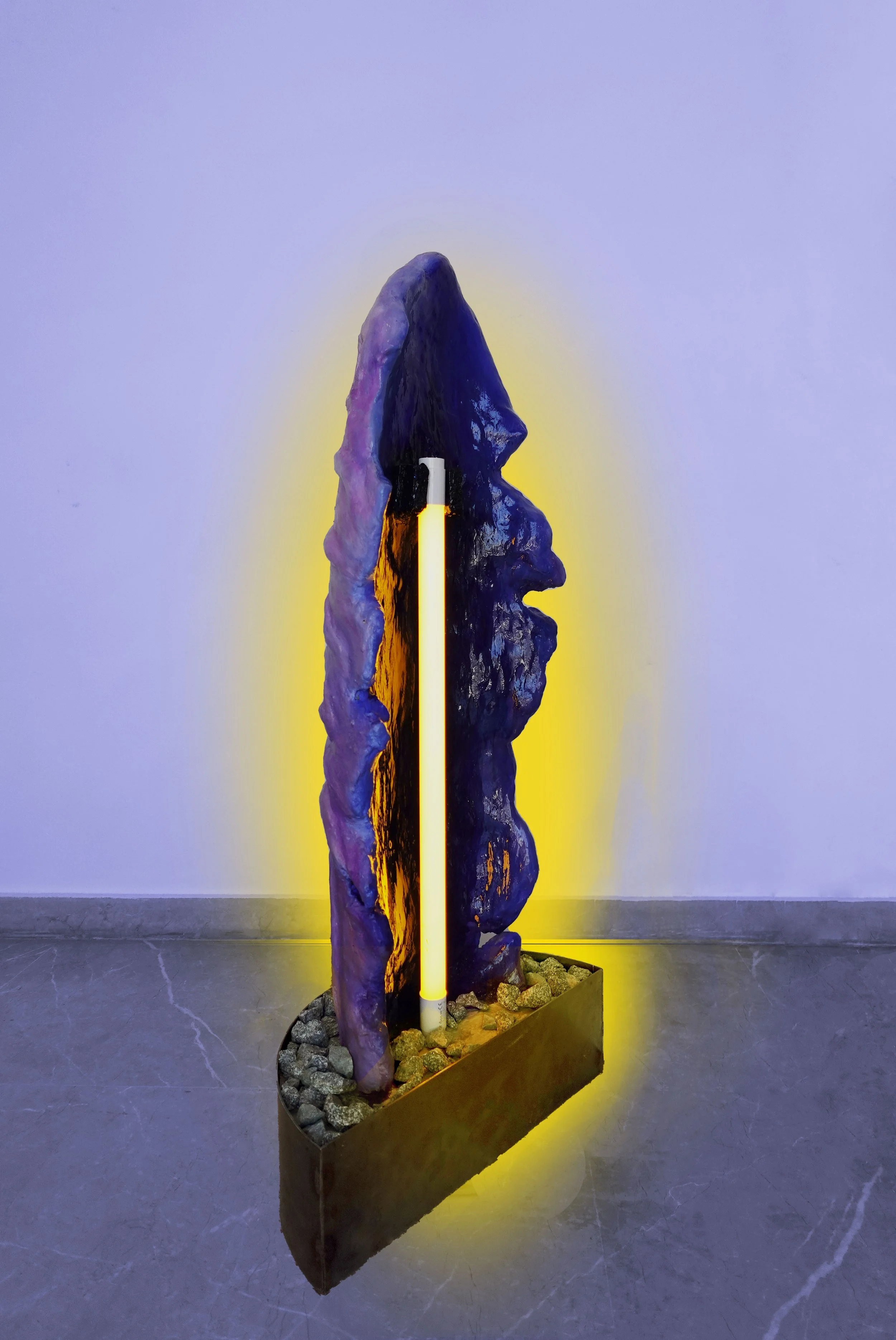 Art installation featuring a vertically standing sculpture that resembles a human face profile, illuminated by yellow and purple lighting, with rocks at the base, placed against a plain white wall.