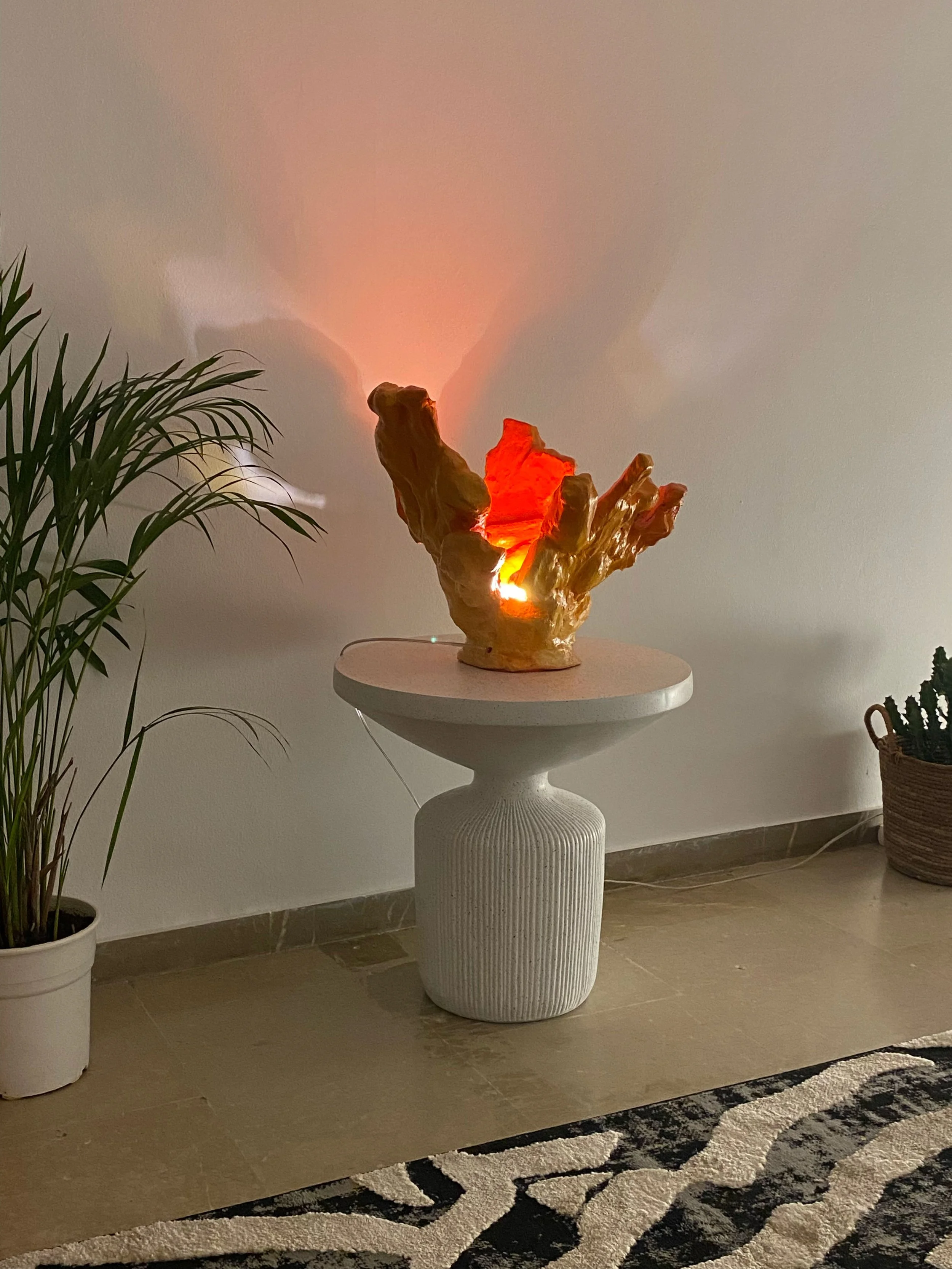 Decorative lamp on a white pedestal table illuminating a room with a warm glow, with potted plants on either side and a patterned rug on the floor.