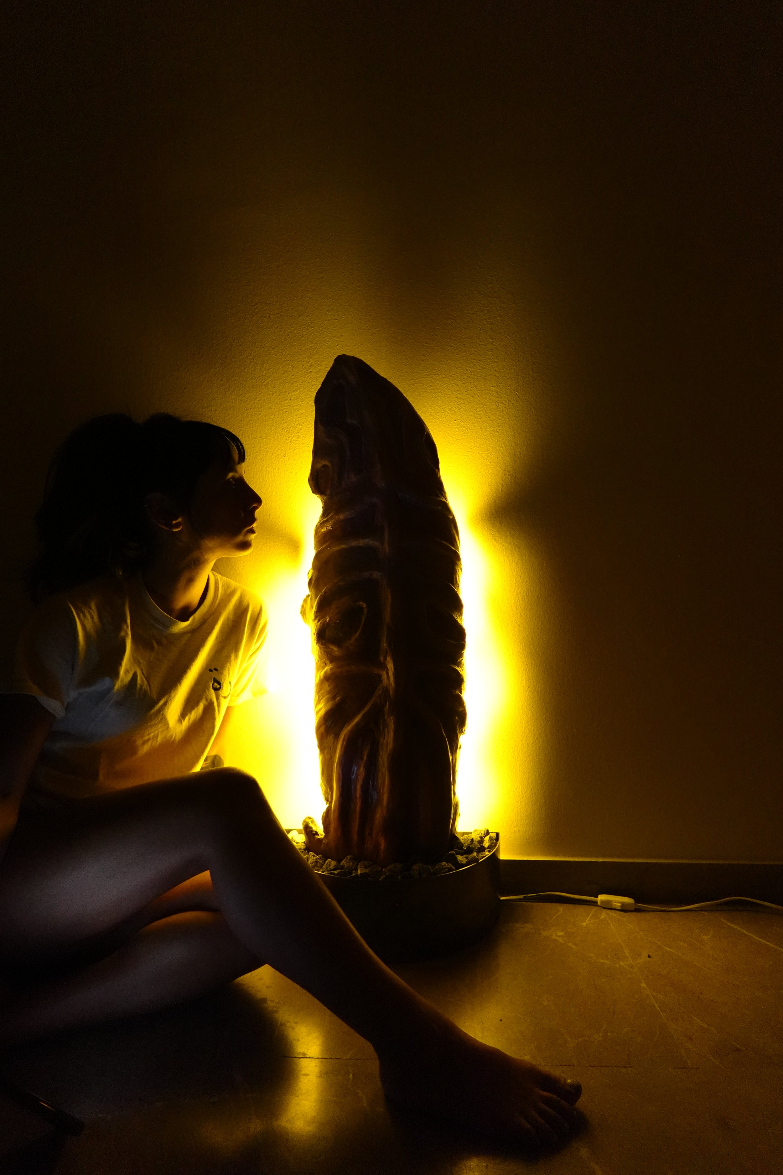 Woman sitting on the floor in front of a lit sculpture resembling a large mushroom or plant, with a yellow glow illuminating the sculpture from behind, and a wall socket and extension cord visible on the floor.