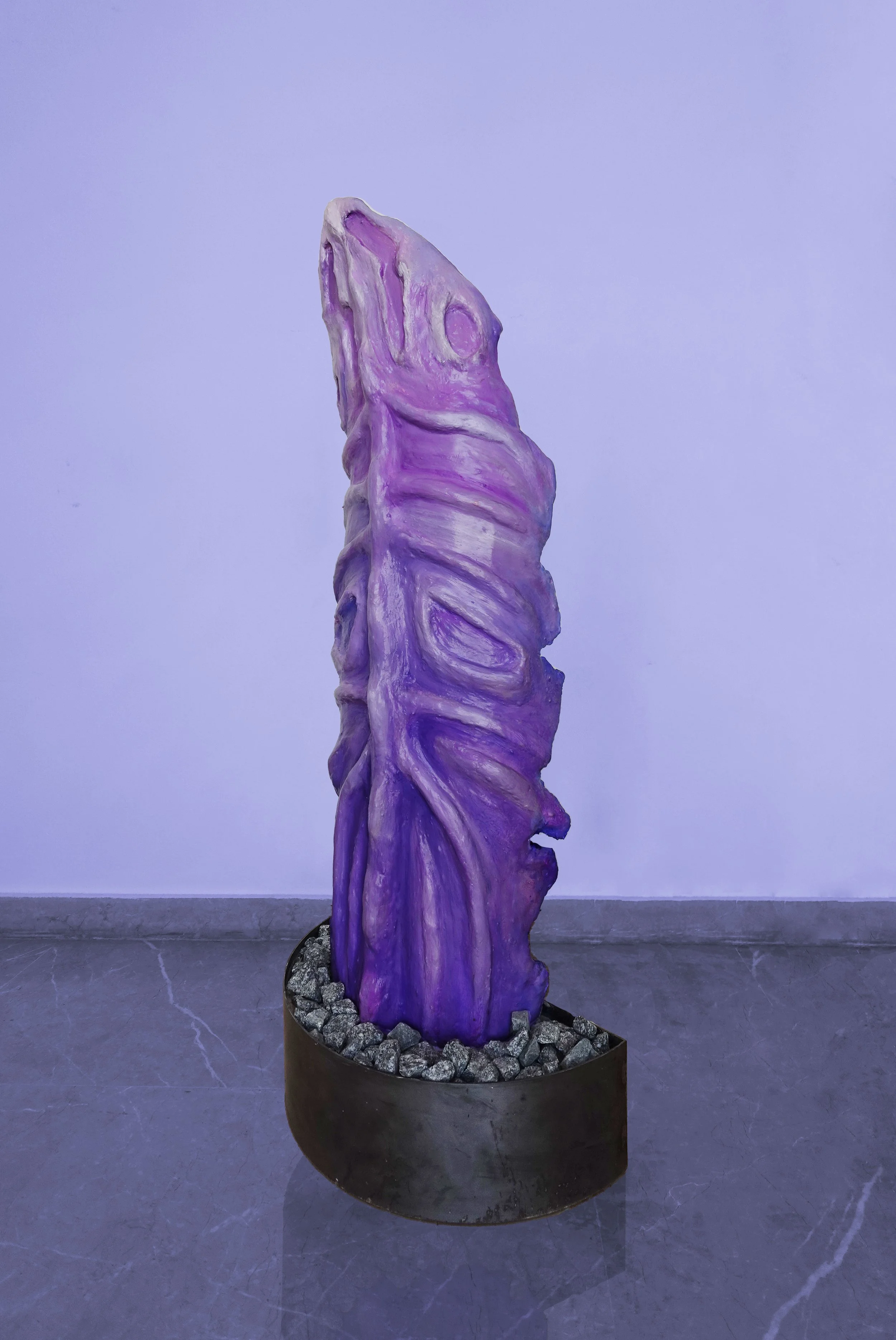 Purple sculpture resembling a rough, elongated shape with textured ridges, displayed on a round base filled with rocks, against a plain background.