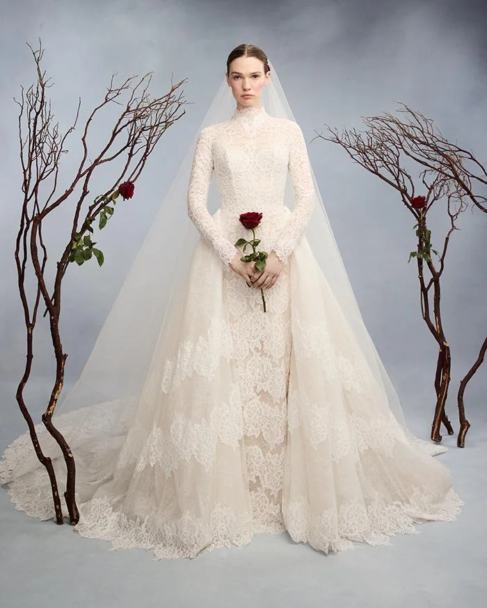 A woman in a white lace wedding gown holding a red rose, standing between three decorative branches with red roses. The background is a cloudy gray sky.