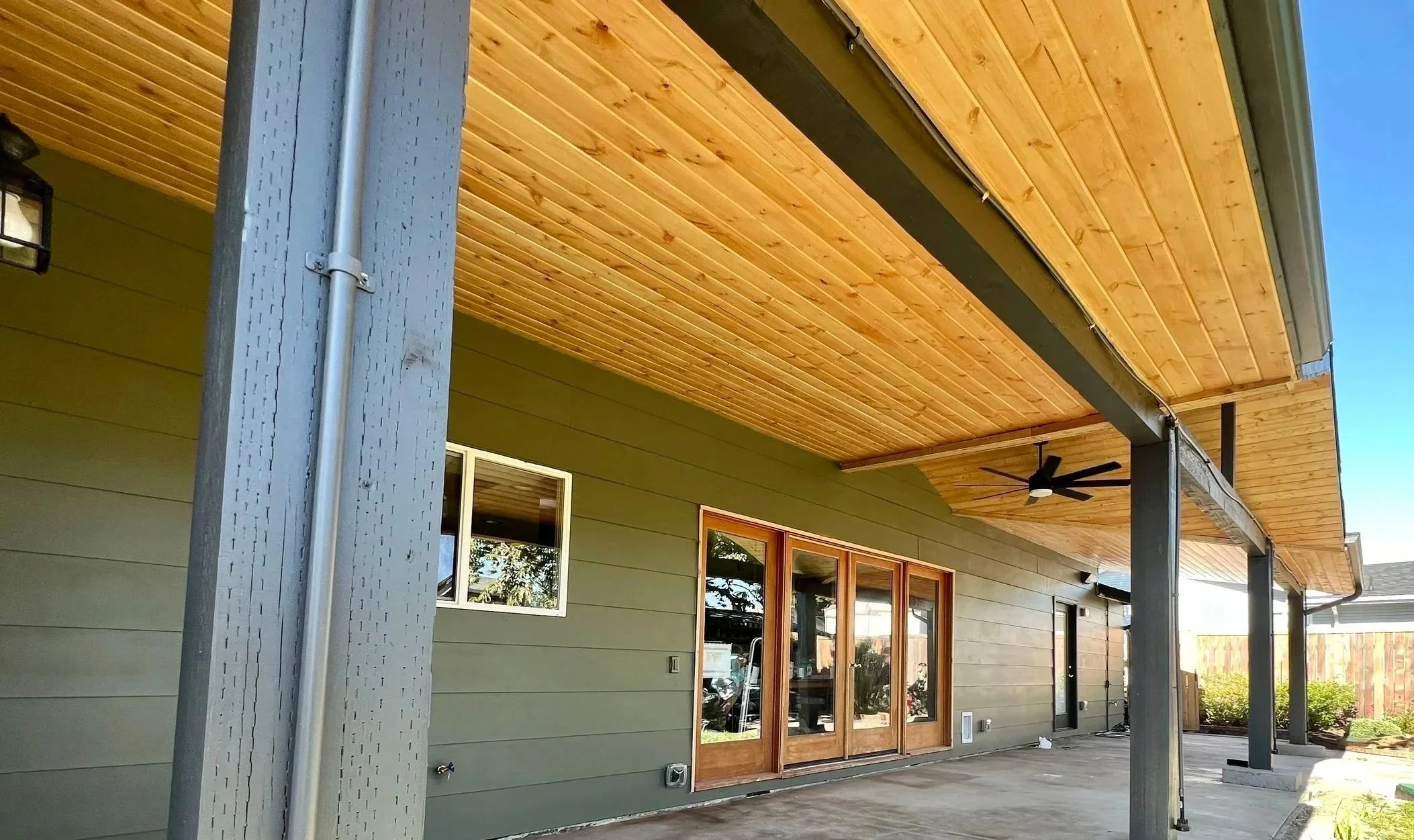 View of a house's outdoor patio area with wooden ceiling, green siding, windows, and a ceiling fan, with a clear sky in the background.