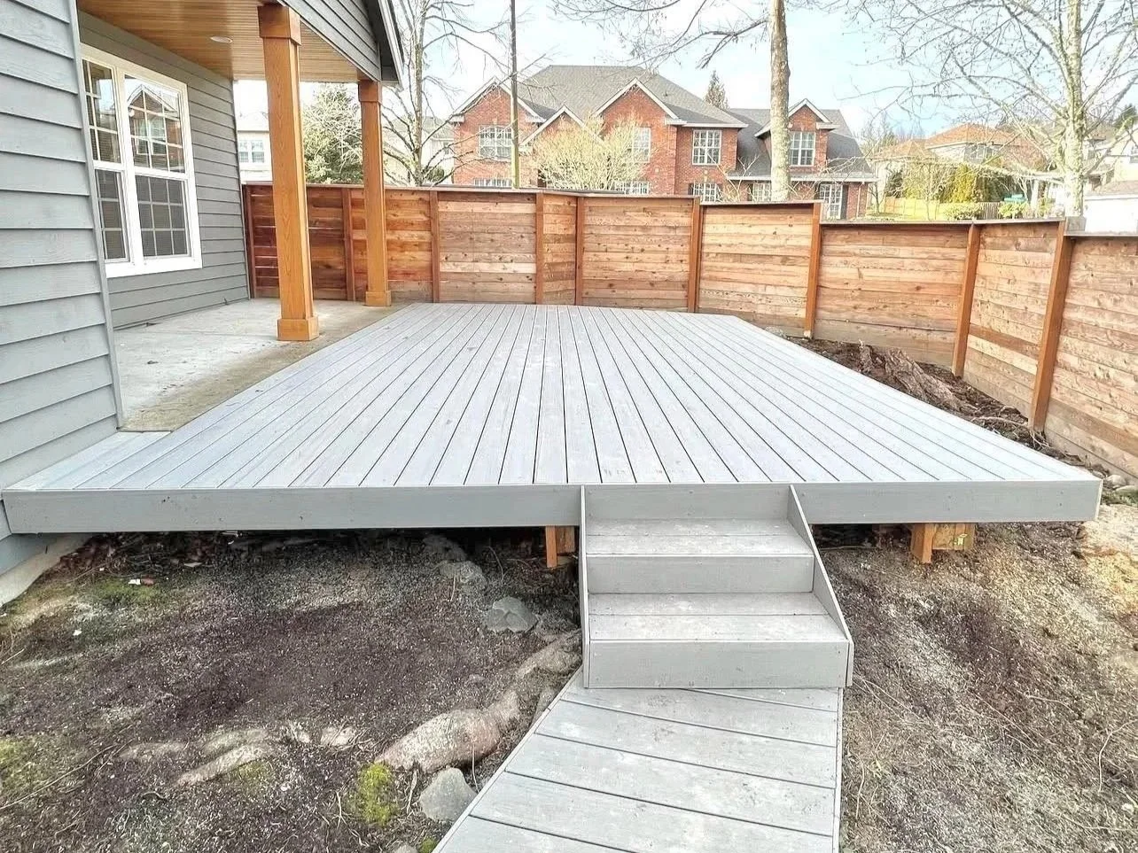 Newly constructed wooden deck with stairs, attached to a house with grey siding and white-framed windows. The deck is surrounded by a wooden privacy fence, with neighboring houses in the background.