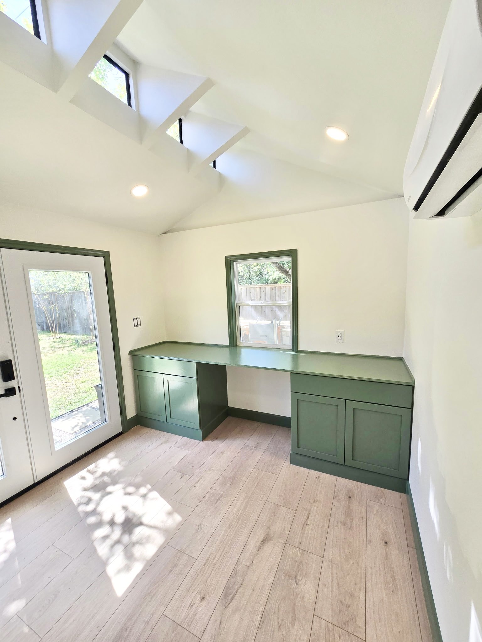 A bright room with light-colored wooden floor, green cabinetry along the wall, two windows and a glass door letting in natural light, and ceiling with skylights and recessed lighting.
