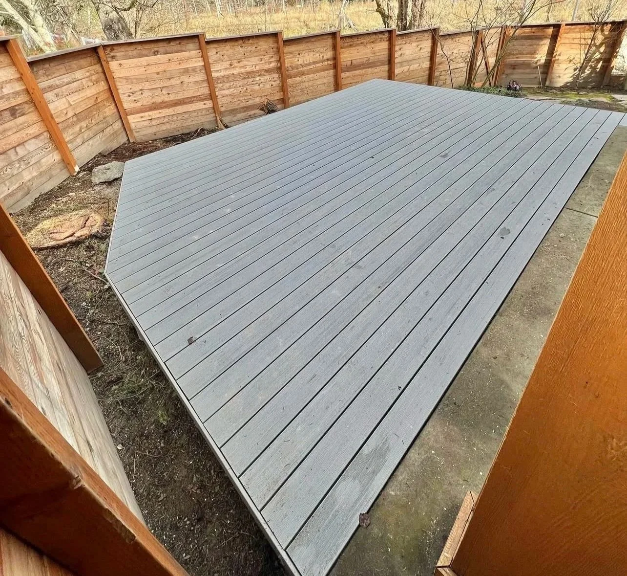 A wooden deck in a backyard enclosed by a wooden fence, with some dirt and rocks visible on the ground surrounding the deck.