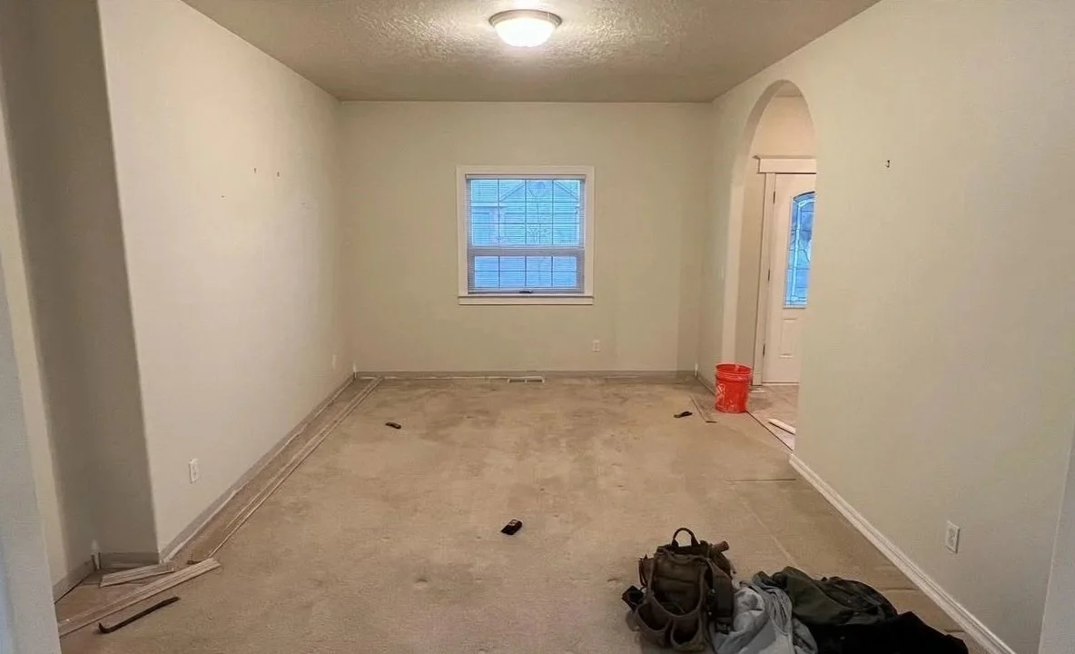 Empty living room with light-colored walls, carpeted floor, window on the far wall, and an archway door to another room. Construction tools and a backpack are on the floor.