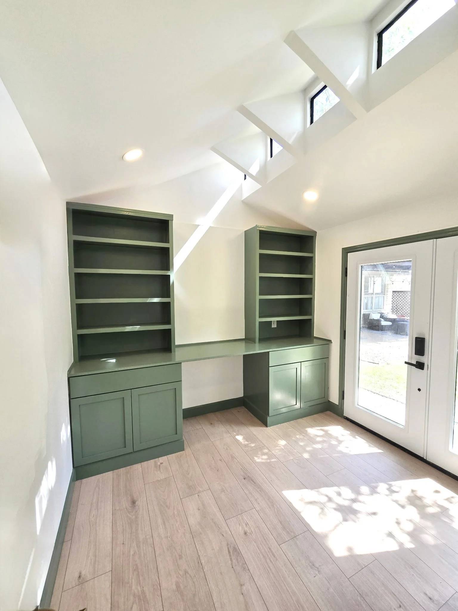 Interior of a room with light wooden flooring, green built-in shelves and cabinets, and a glass door leading outside. Sunlight streams through multiple skylight windows in the ceiling.